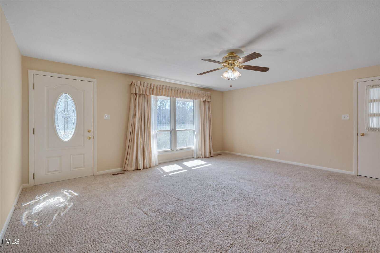 Empty beige carpeted room with a ceiling fan, two doors, a large window with drapes letting in sunlight, and white trim along the walls. The room appears clean and well-lit.