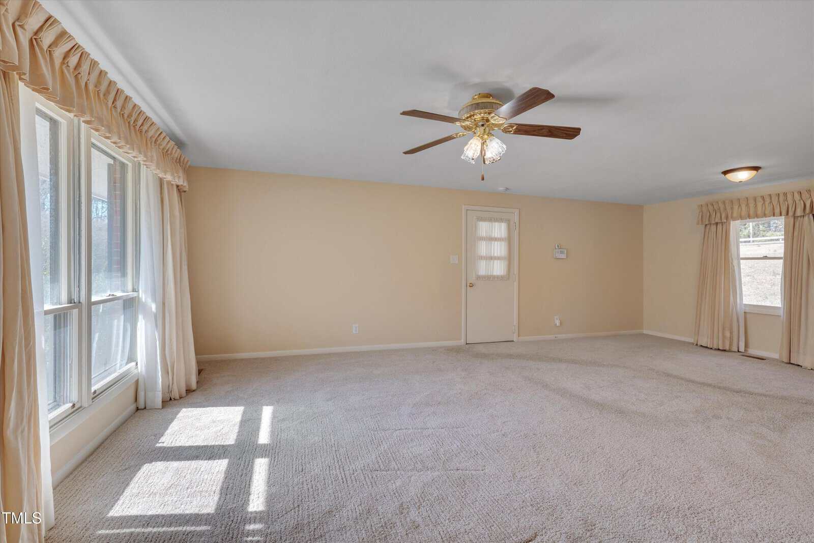 A spacious, carpeted living room with beige walls, large windows with drapes, a ceiling fan with lights, and a door leading outside. Sunlight streams through the windows, creating rectangular patterns on the floor.