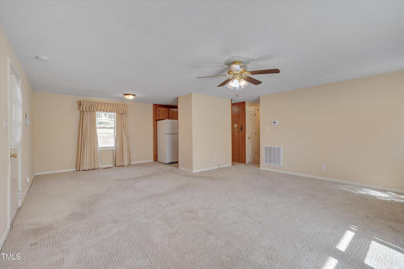 A beige carpeted living room with cream walls, a ceiling fan with lights, a window with curtains, and a partial view of a white refrigerator and kitchen area in the background. Sunlight streams through a window.