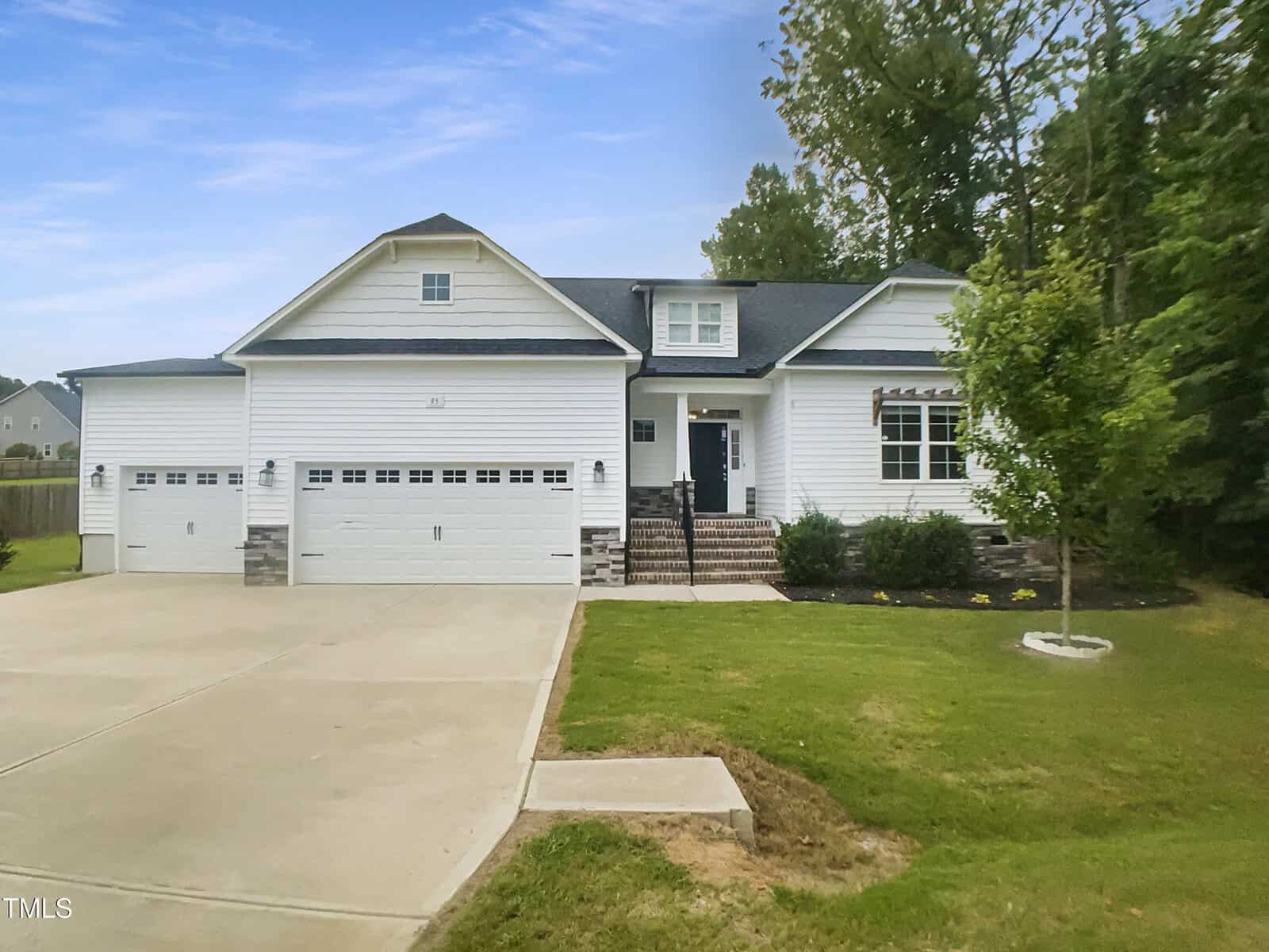 A white single-story house with a gray roof, three-car garage, brick steps leading to the front door, and a neatly maintained front yard with a small tree and surrounding greenery.