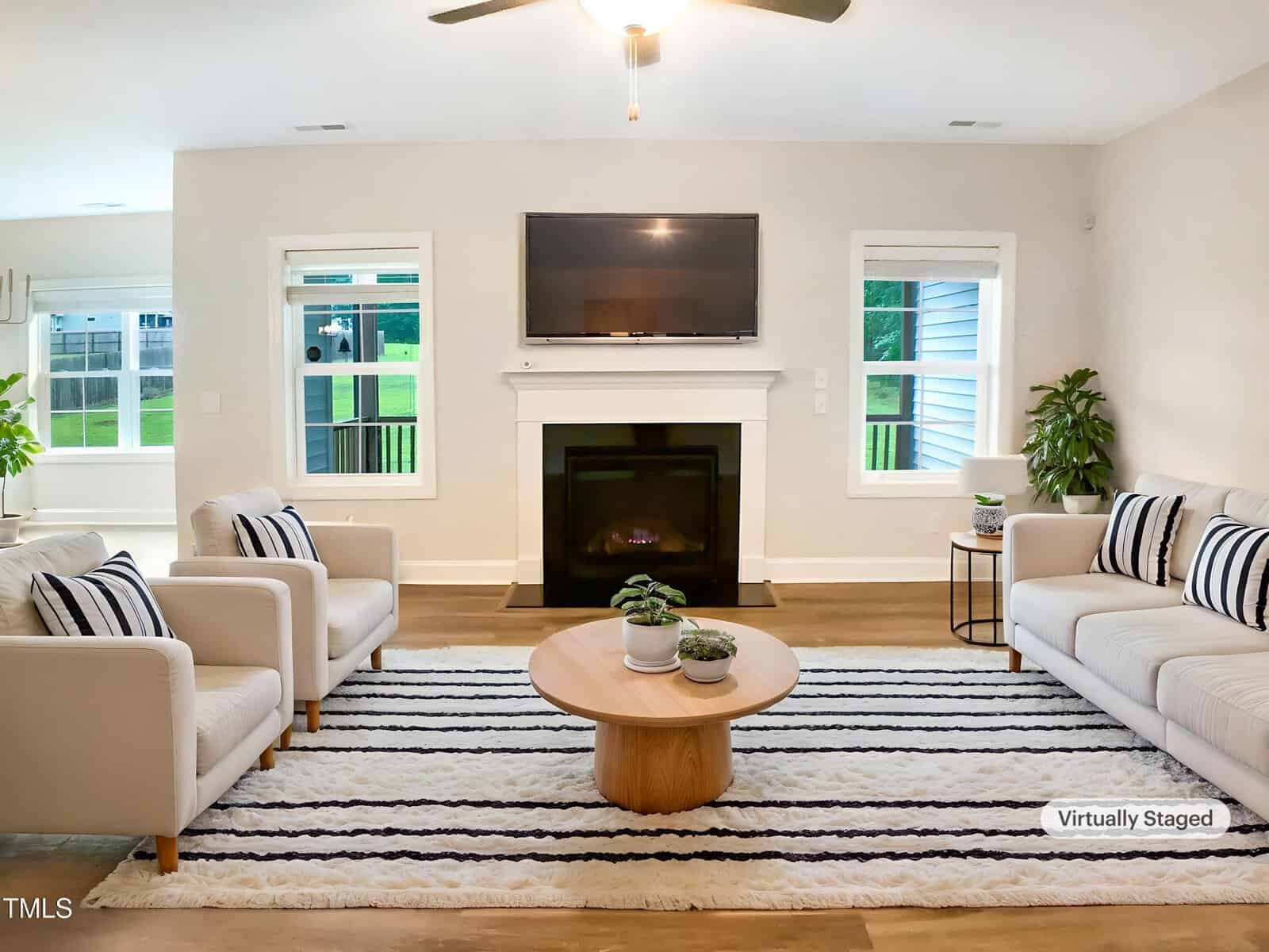 A modern living room with light-colored walls, a striped rug, a beige sofa, two armchairs, a round wooden coffee table, potted plants, and a fireplace with a wall-mounted TV above it. The image is virtually staged.