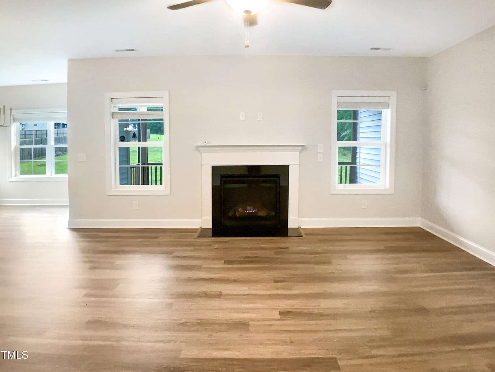 A living room with light-colored walls, two windows flanking a central white fireplace, wood flooring, and a ceiling fan. There is an open view into another room on the left side.