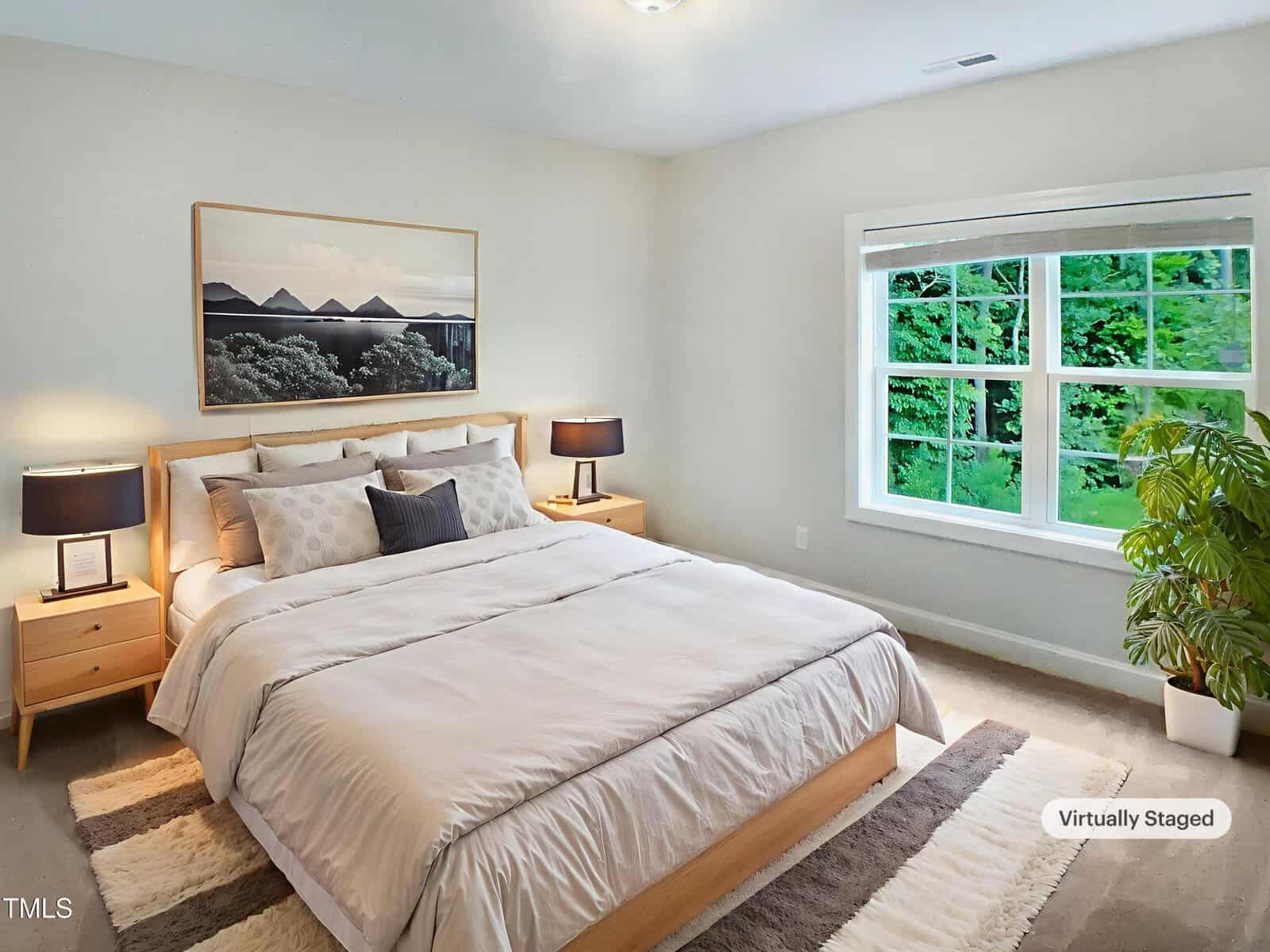 A bright bedroom with a large bed, light wood frame, two nightstands with black lamps, a potted plant, a large window with greenery outside, and a landscape picture on the wall; labeled as virtually staged.