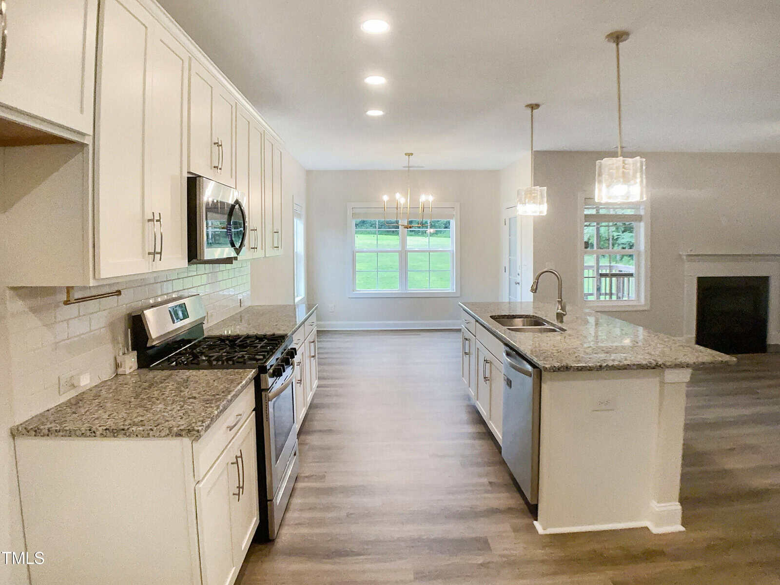 Modern kitchen with white cabinets, stainless steel appliances, granite countertops, and a center island. Pendant lights hang over the island, and large windows in the dining area provide natural light. Wood flooring throughout.