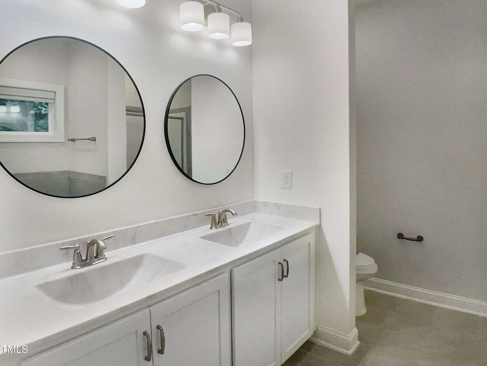 Modern bathroom with double sinks, two round mirrors, white cabinets, marble countertop, silver faucets, overhead lights, and a toilet visible in a separate area. A window is reflected in one mirror.