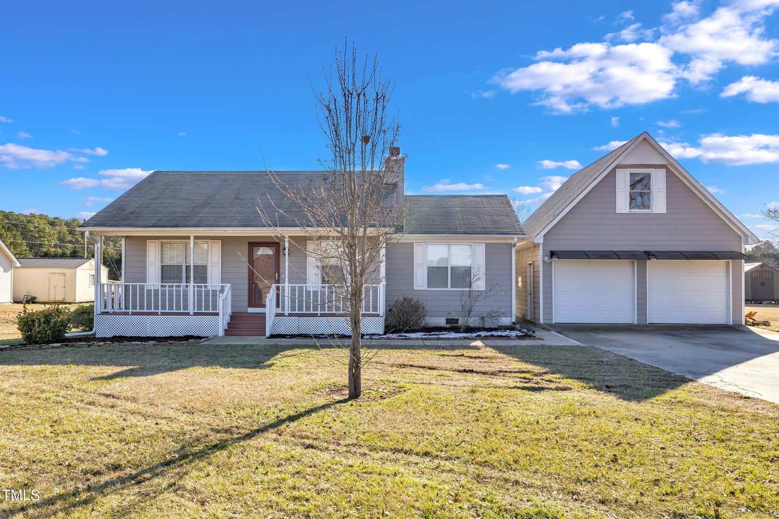 A single-story beige house with a covered front porch, a leafless tree in the yard, and an attached two-car garage with an upper-level dormer, set on a sunny day with a blue sky and scattered clouds.