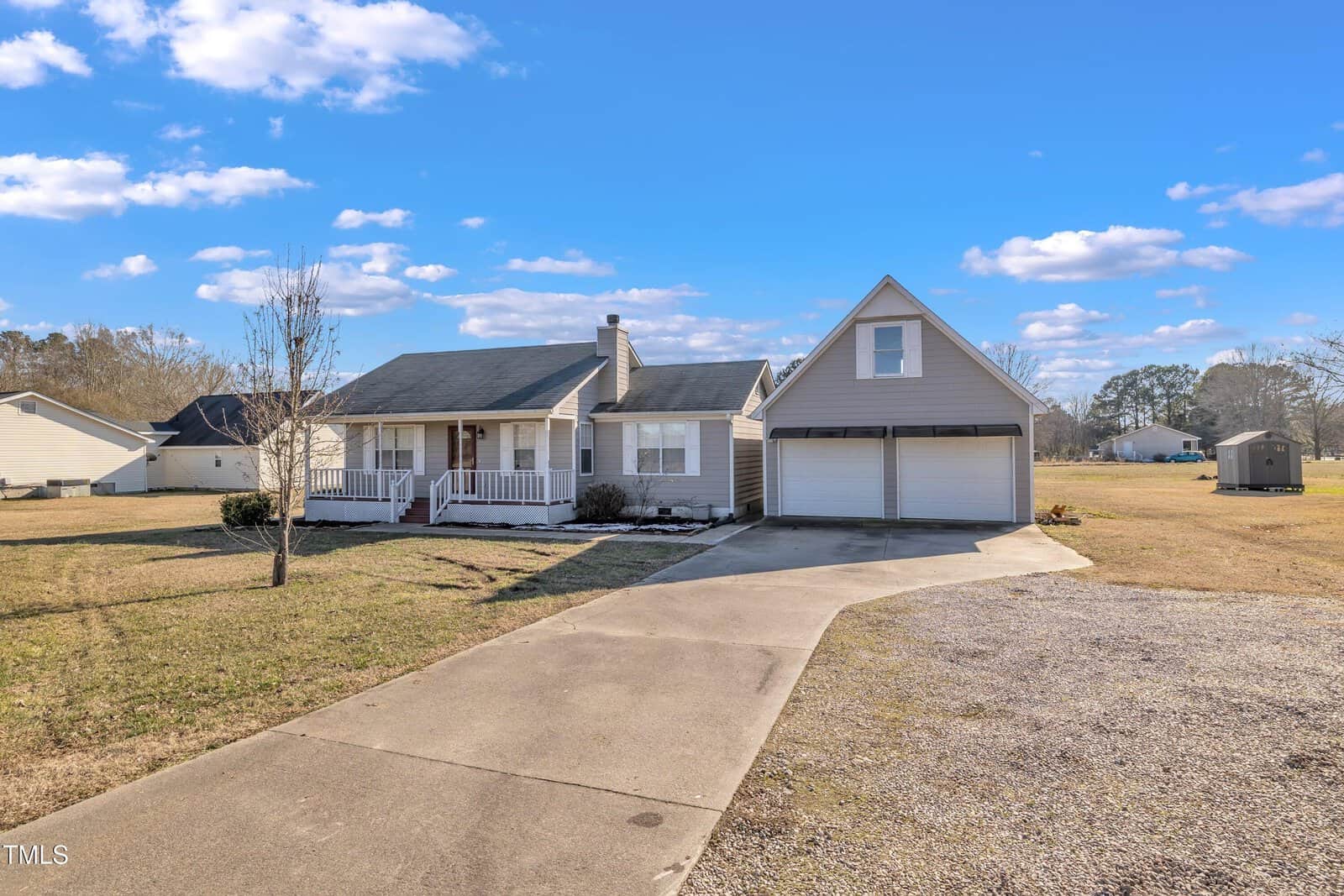 Single-story light gray house with a covered front porch, detached two-car garage with an upper level, concrete driveway, and spacious yard under a blue sky with scattered clouds.
