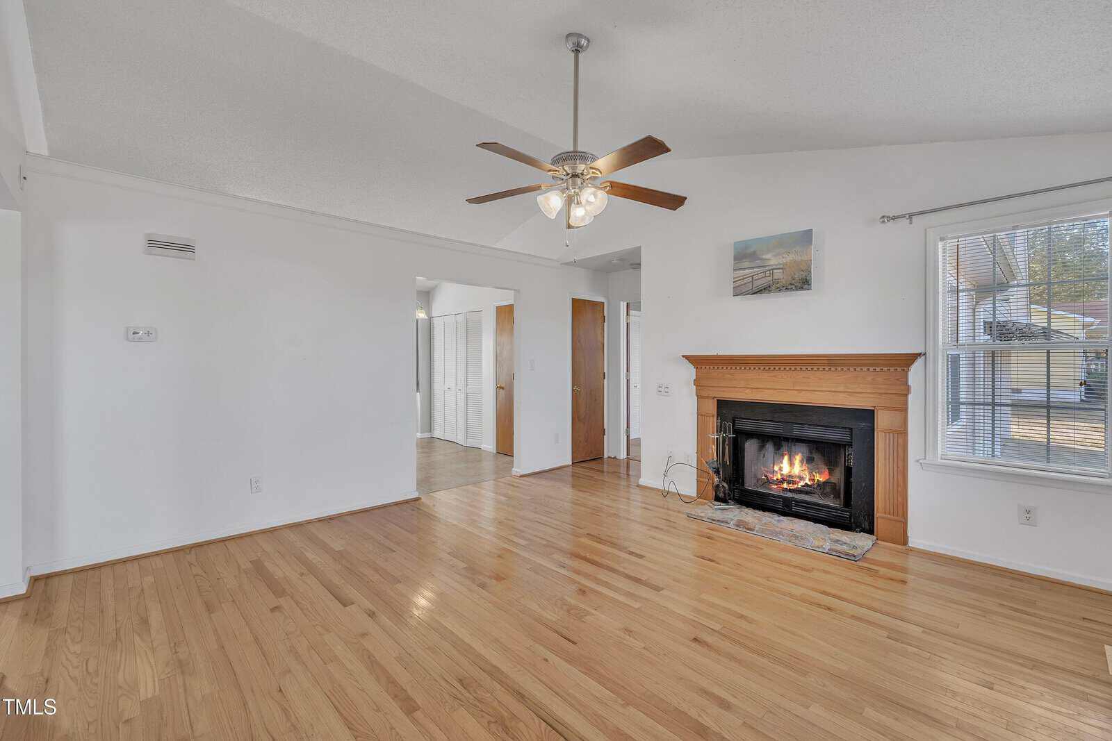 A living room with light wood floors, white walls, a ceiling fan with lights, and a lit fireplace with a wooden mantel. A window lets in sunlight, and there are doorways leading to other rooms.