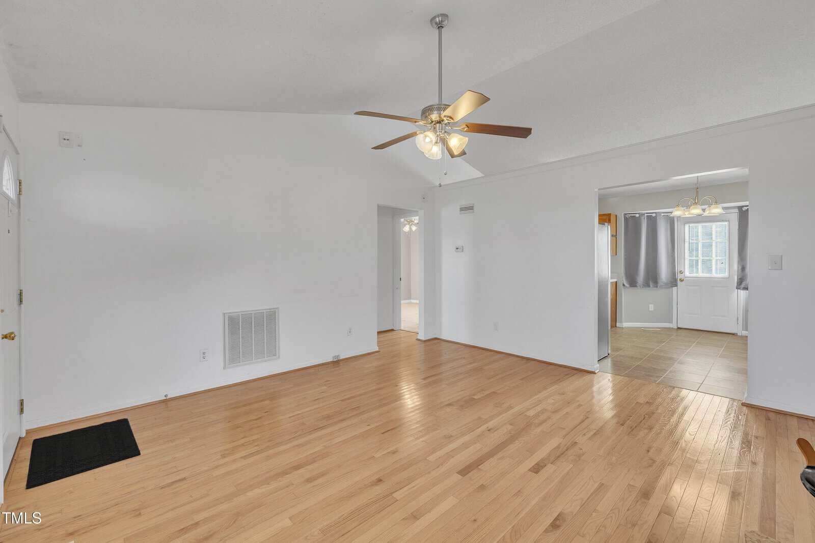 A room with light-colored hardwood floors, white walls, a ceiling fan with lights, and a view into an adjacent tiled dining area with a window and chandelier. The space is unfurnished and well-lit.