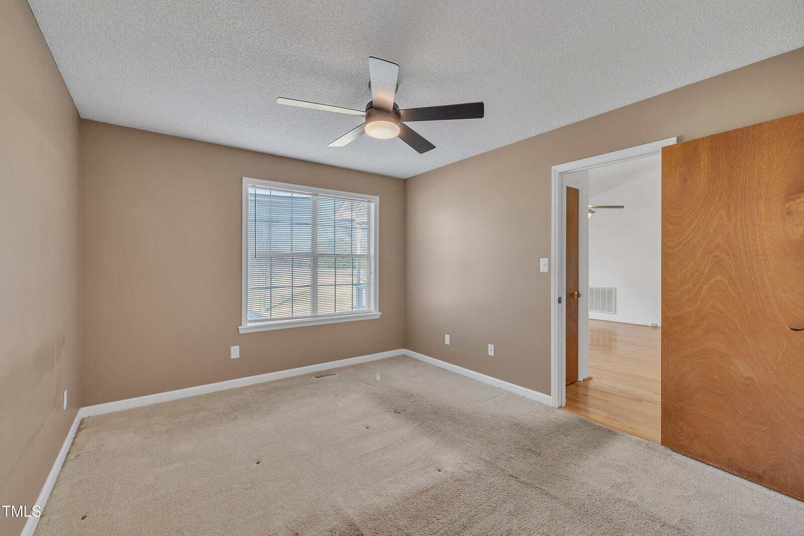 Empty bedroom with beige walls, a single large window with blinds, beige carpet, a ceiling fan with a light, and an open door showing a glimpse of an adjacent room with hardwood floors.