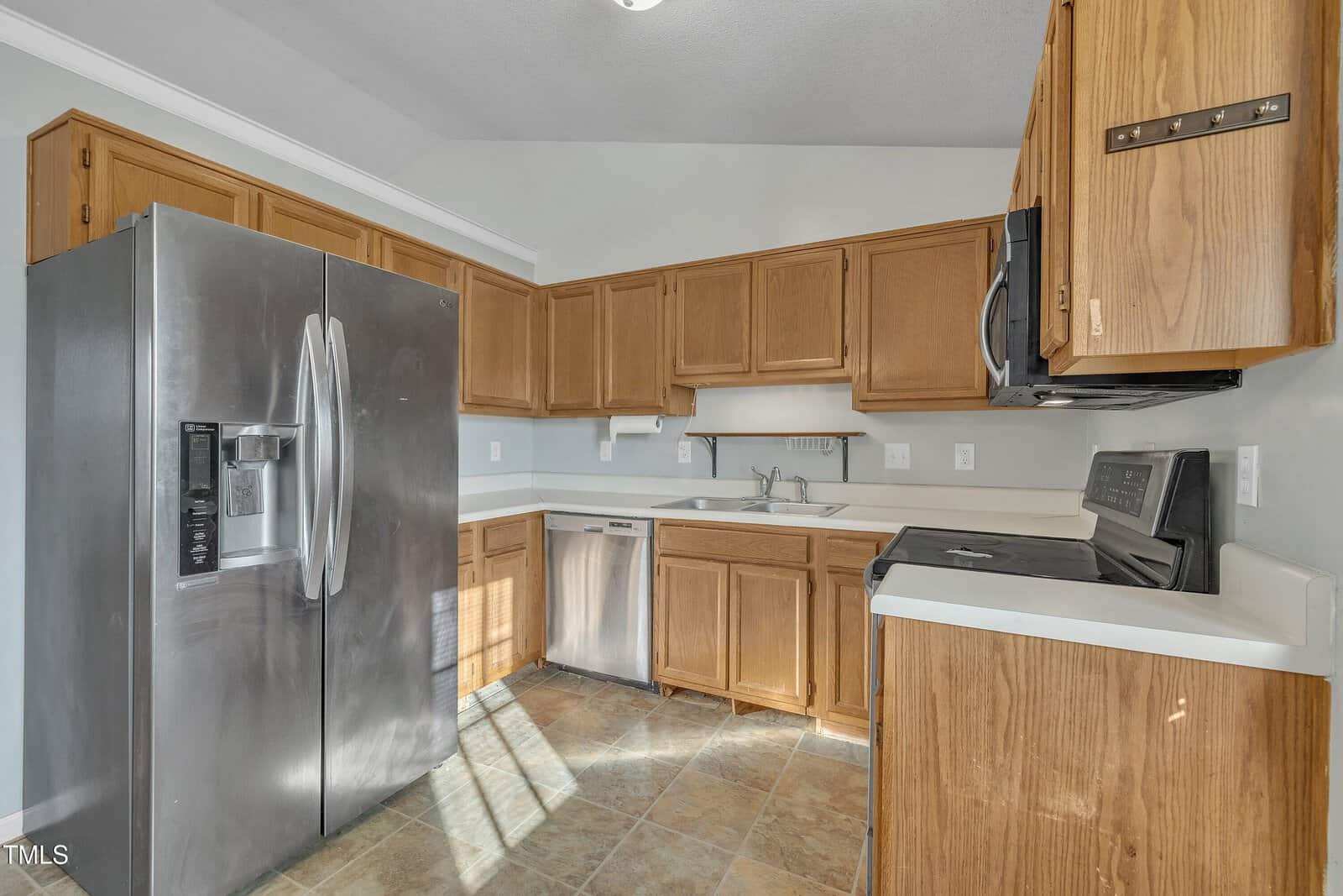 Kitchen with stainless steel refrigerator, dishwasher, oven, microwave, double sink, light wood cabinets, and tile floor. Walls are painted light gray, and countertops are white. Room is well-lit with natural light.