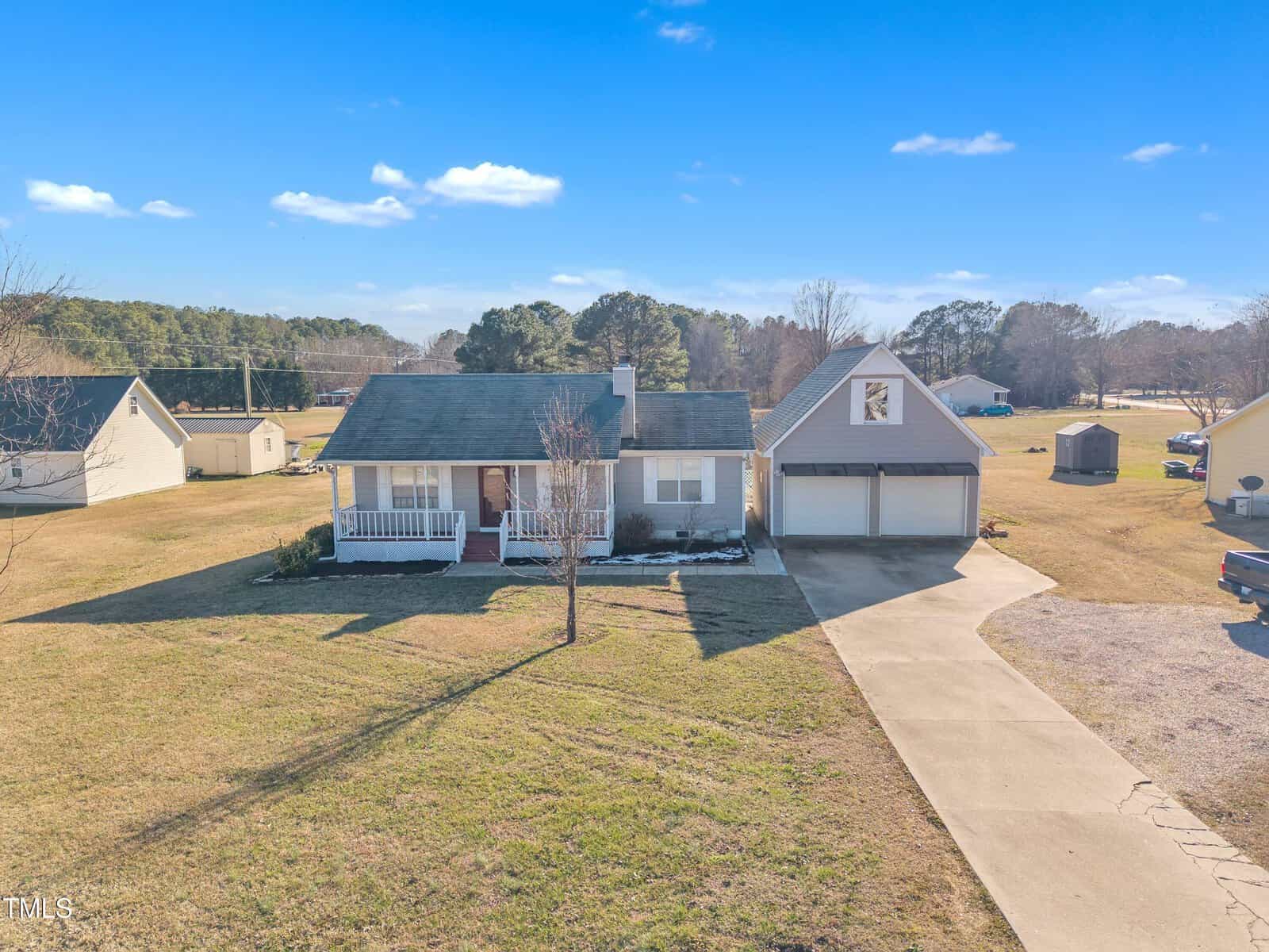 A single-story house with a covered front porch and attached two-car garage, set on a large grassy lot with a paved driveway. There are trees and additional buildings in the background under a clear blue sky.