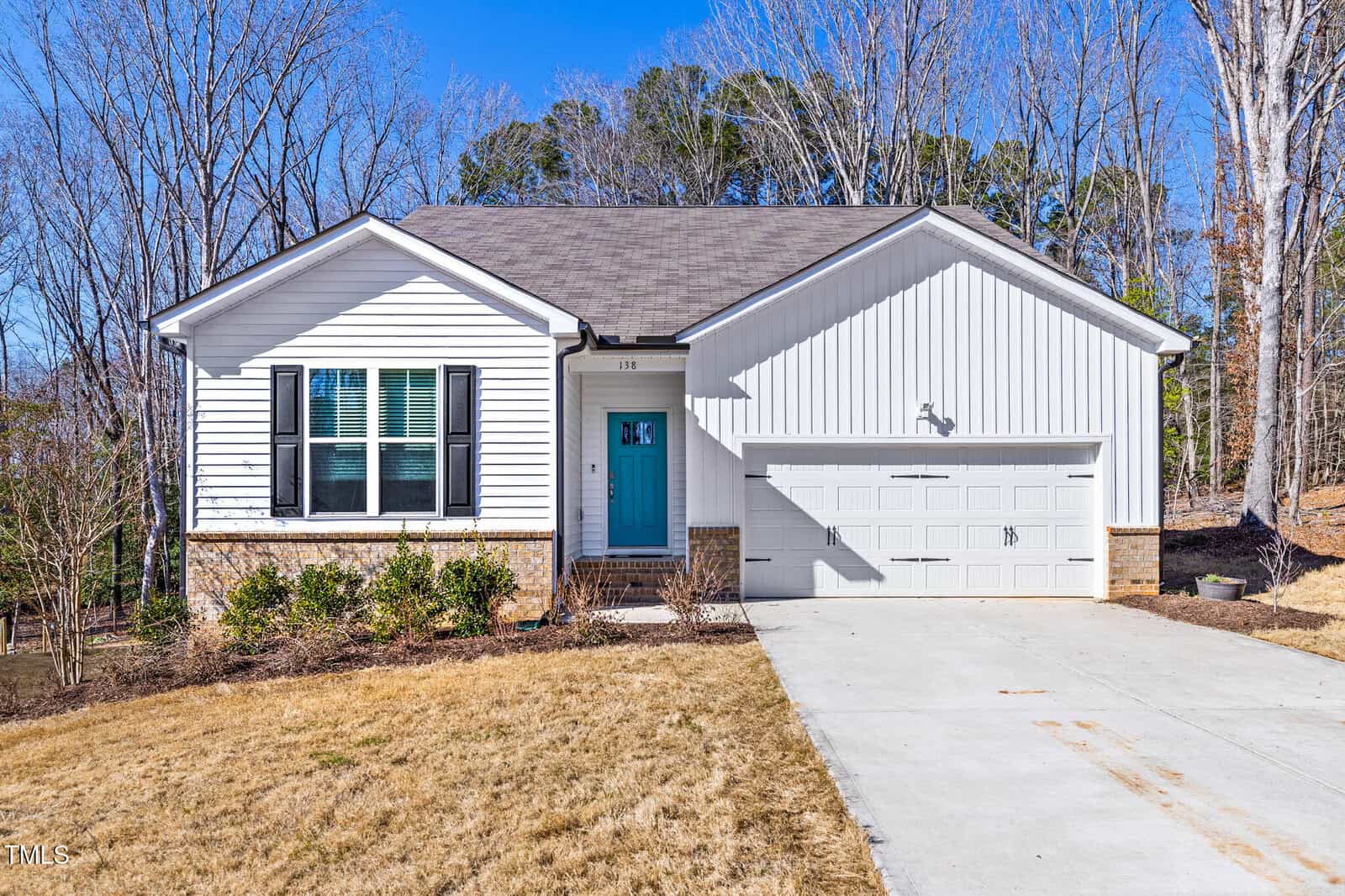 A single-story white house with a blue front door, attached two-car garage, concrete driveway, brick foundation, and a small front yard with minimal landscaping. Leafless trees and blue sky in the background.