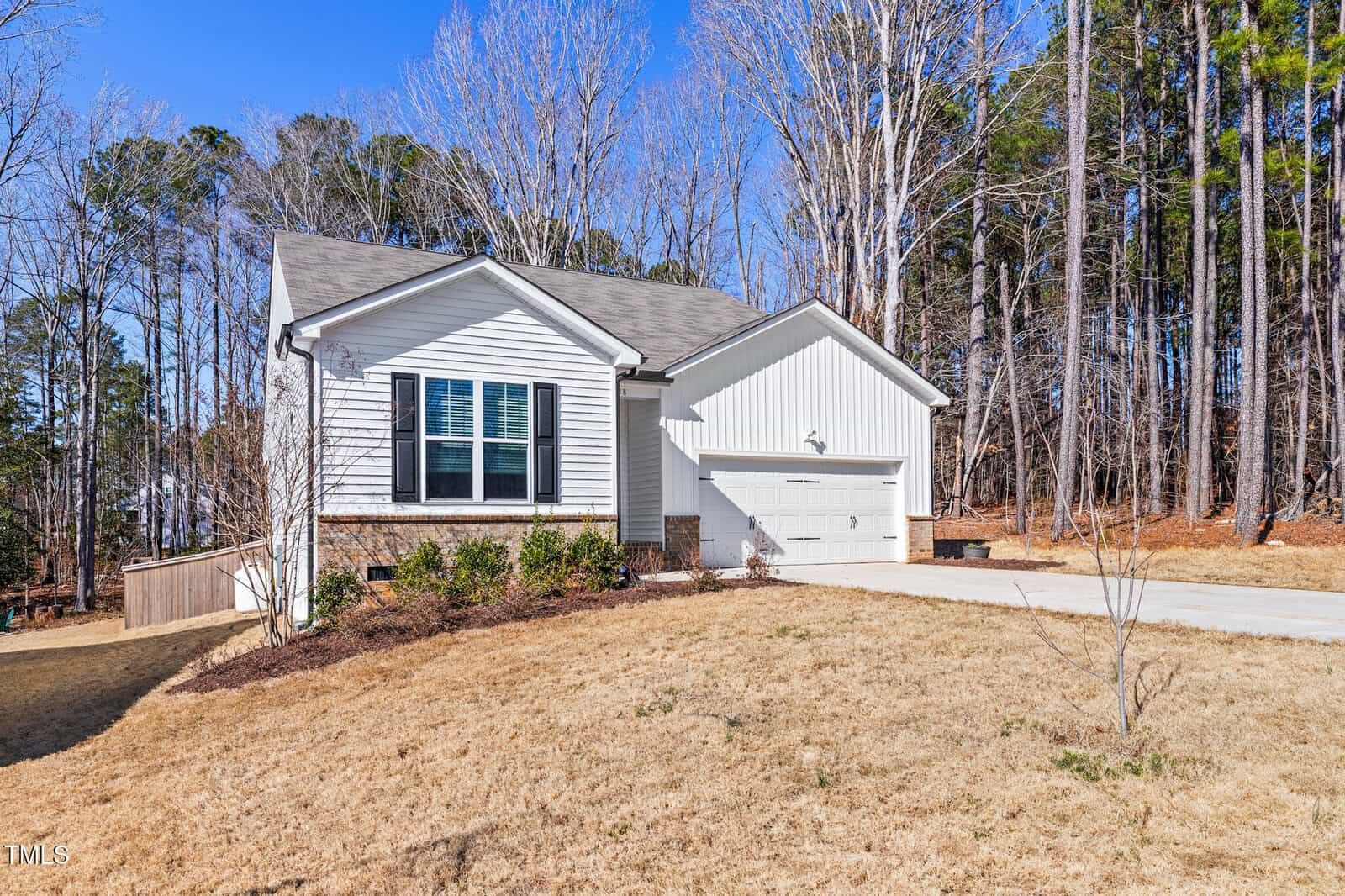 Single-story white house with attached two-car garage, front porch, and large windows. The yard has dry grass and minimal landscaping. Tall trees and a forested area are in the background under a clear blue sky.