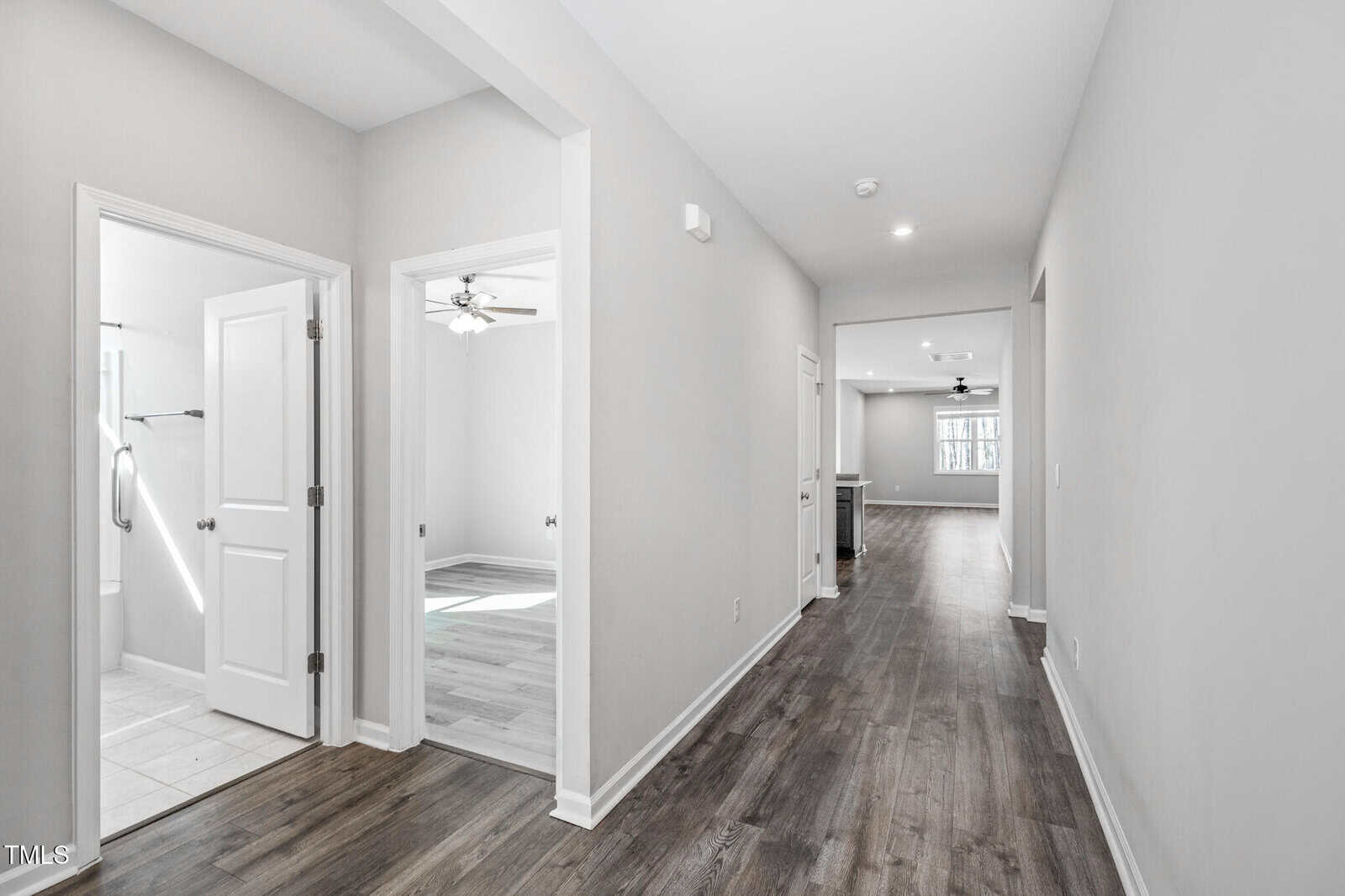 A hallway with wood flooring leads to an open living area. Two doorways on the left reveal a bathroom with tile flooring and a room with a ceiling fan and natural light. Walls are painted light gray.