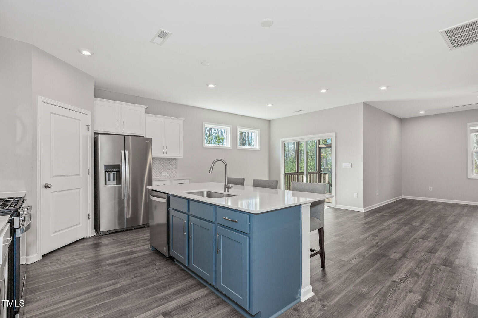 A modern kitchen with stainless steel appliances, white cabinets, a blue island with a sink, gray chairs, light gray walls, large windows, and wood-style flooring, opening to a spacious living area.