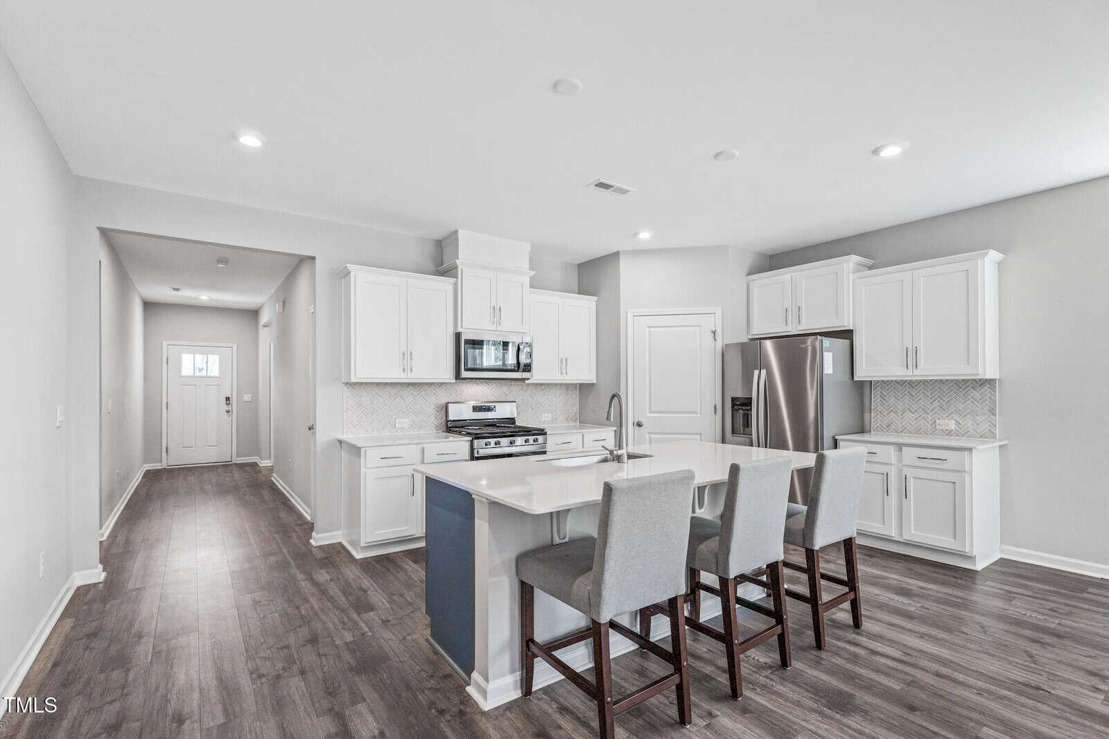 Modern kitchen with white cabinets, stainless steel appliances, a center island with sink, and three upholstered bar stools. The kitchen opens to a hallway with wood flooring and a door visible at the far end.