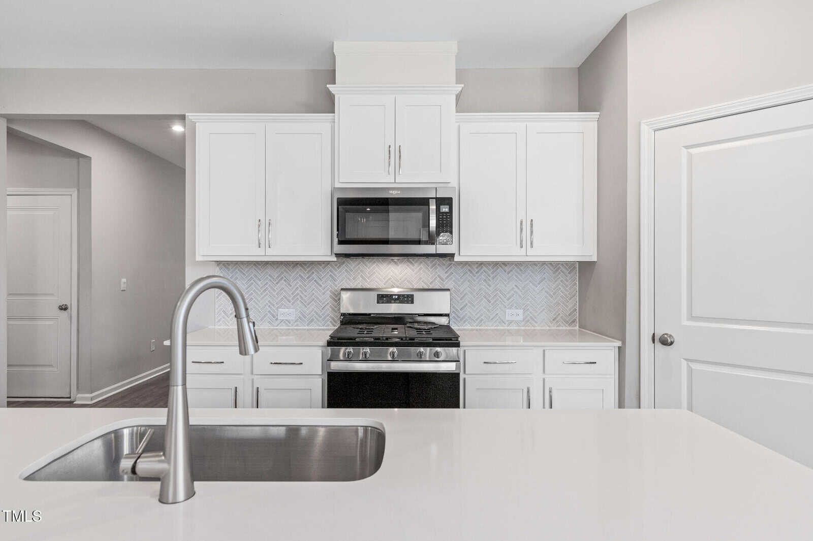 Modern kitchen with white cabinets, a stainless steel oven and microwave, a herringbone tile backsplash, and a white countertop with an integrated sink and faucet in the foreground. Bright, clean, and minimalistic design.
