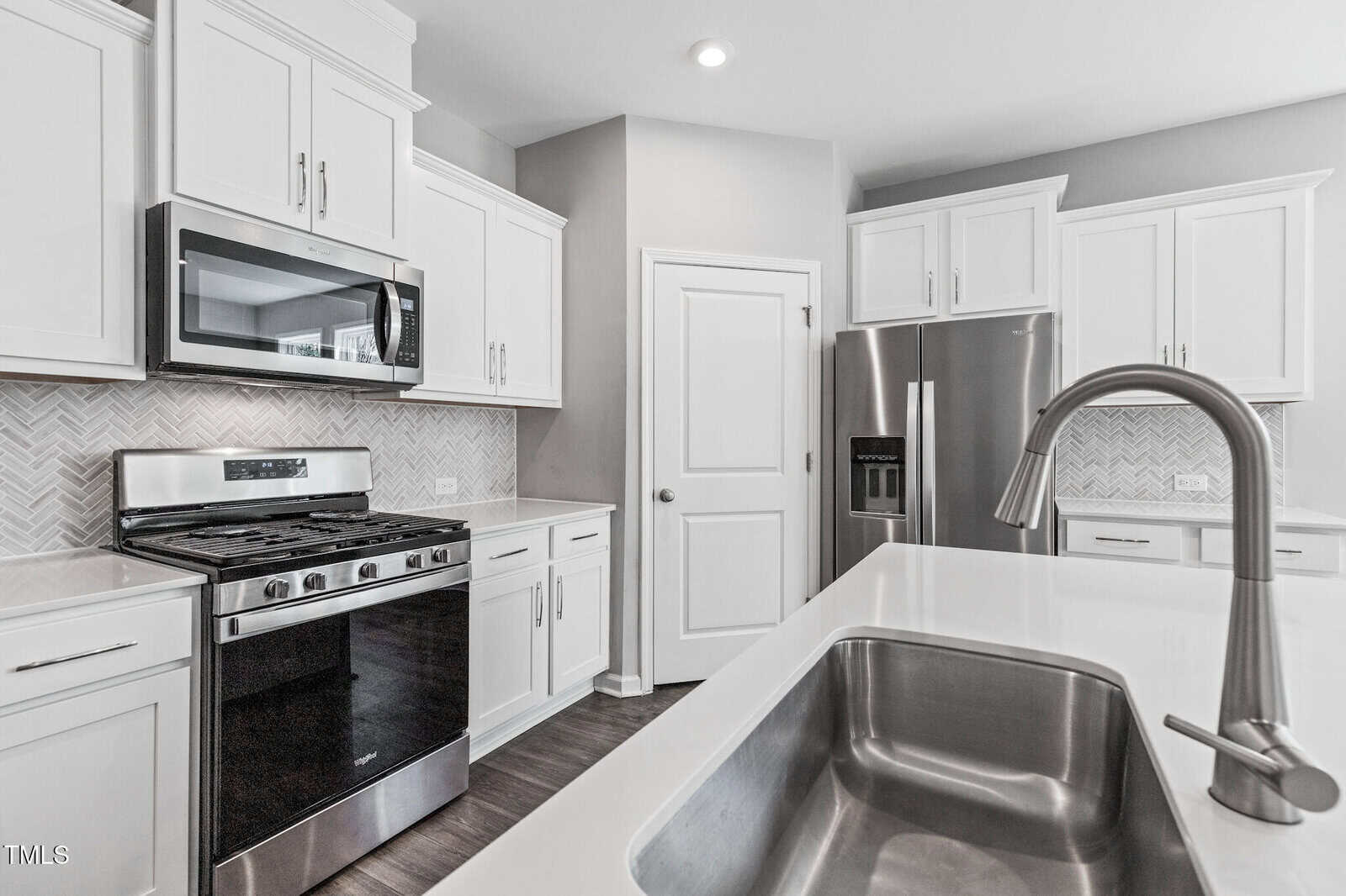 Modern kitchen with white cabinets, stainless steel appliances including a gas stove, microwave, and refrigerator, a central island with a built-in sink and faucet, and a tile backsplash in a herringbone pattern.