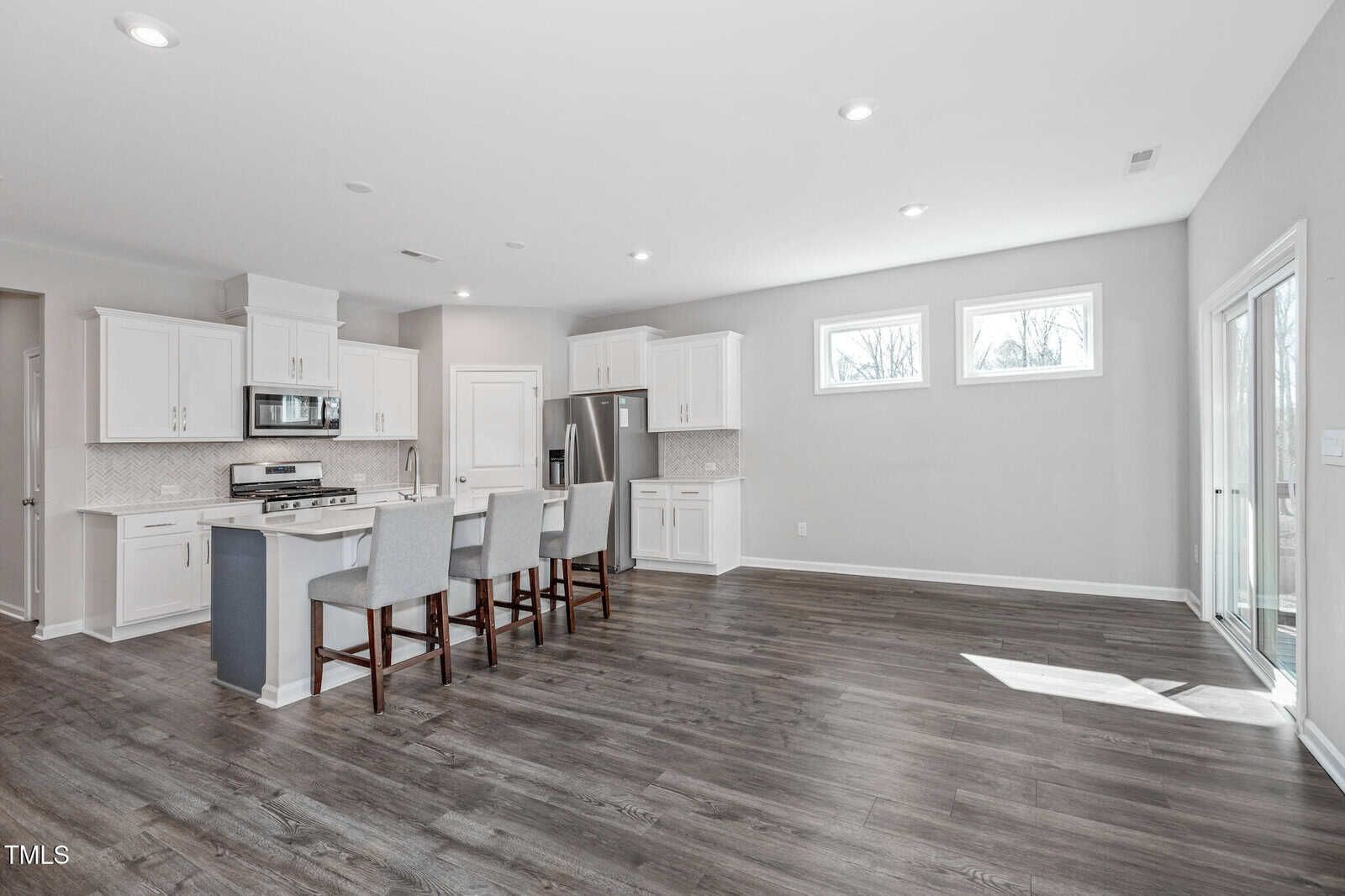 Modern kitchen with white cabinets, stainless steel appliances, island with three gray stools, light wood flooring, and large windows letting in natural light. Sliding glass door on the right opens to the outside.