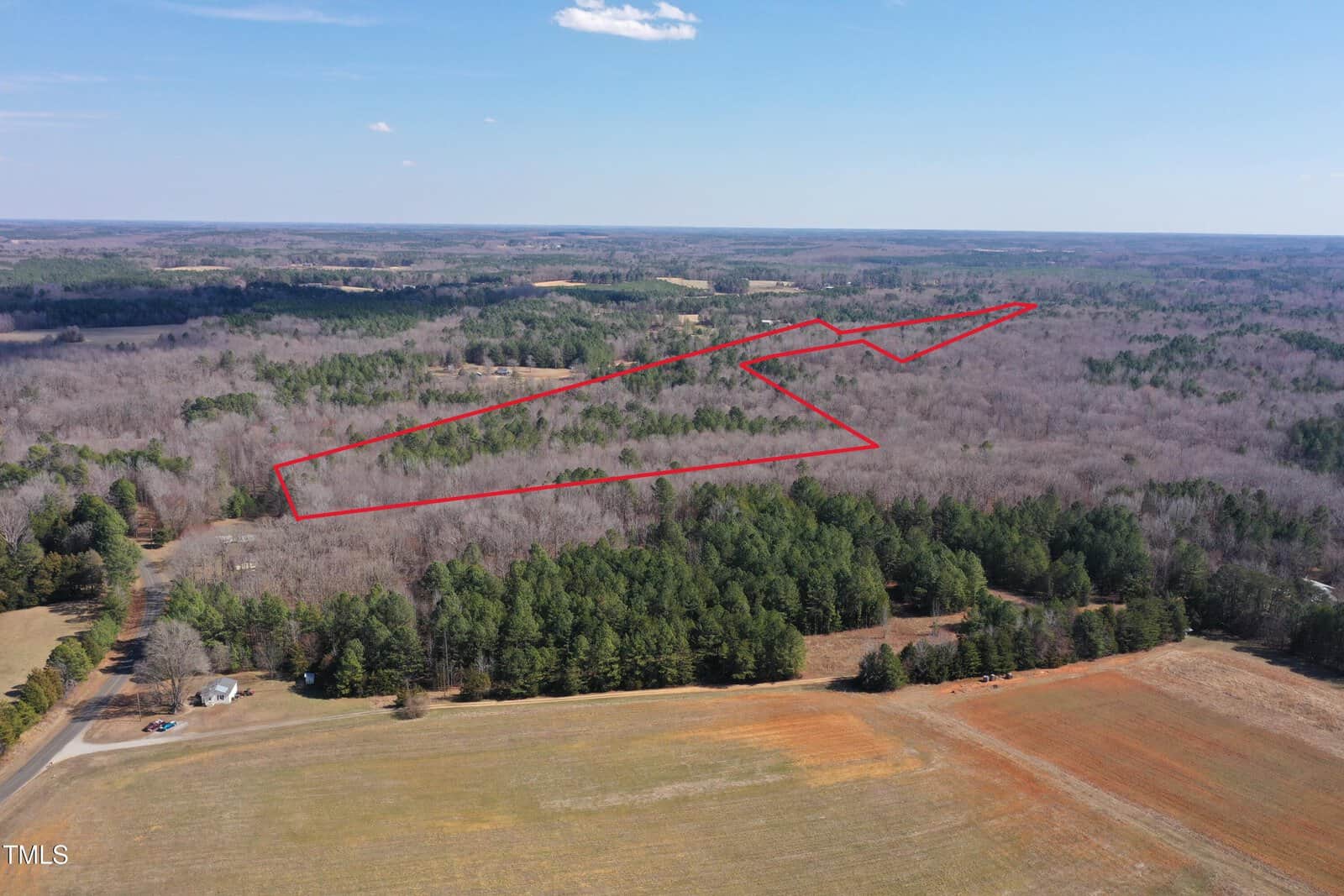 Aerial view of a forested landscape with an outlined area in red. The outlined section stretches across a mix of tree-covered and open land. A road and scattered buildings are visible in the foreground, with expansive woodland in the background.