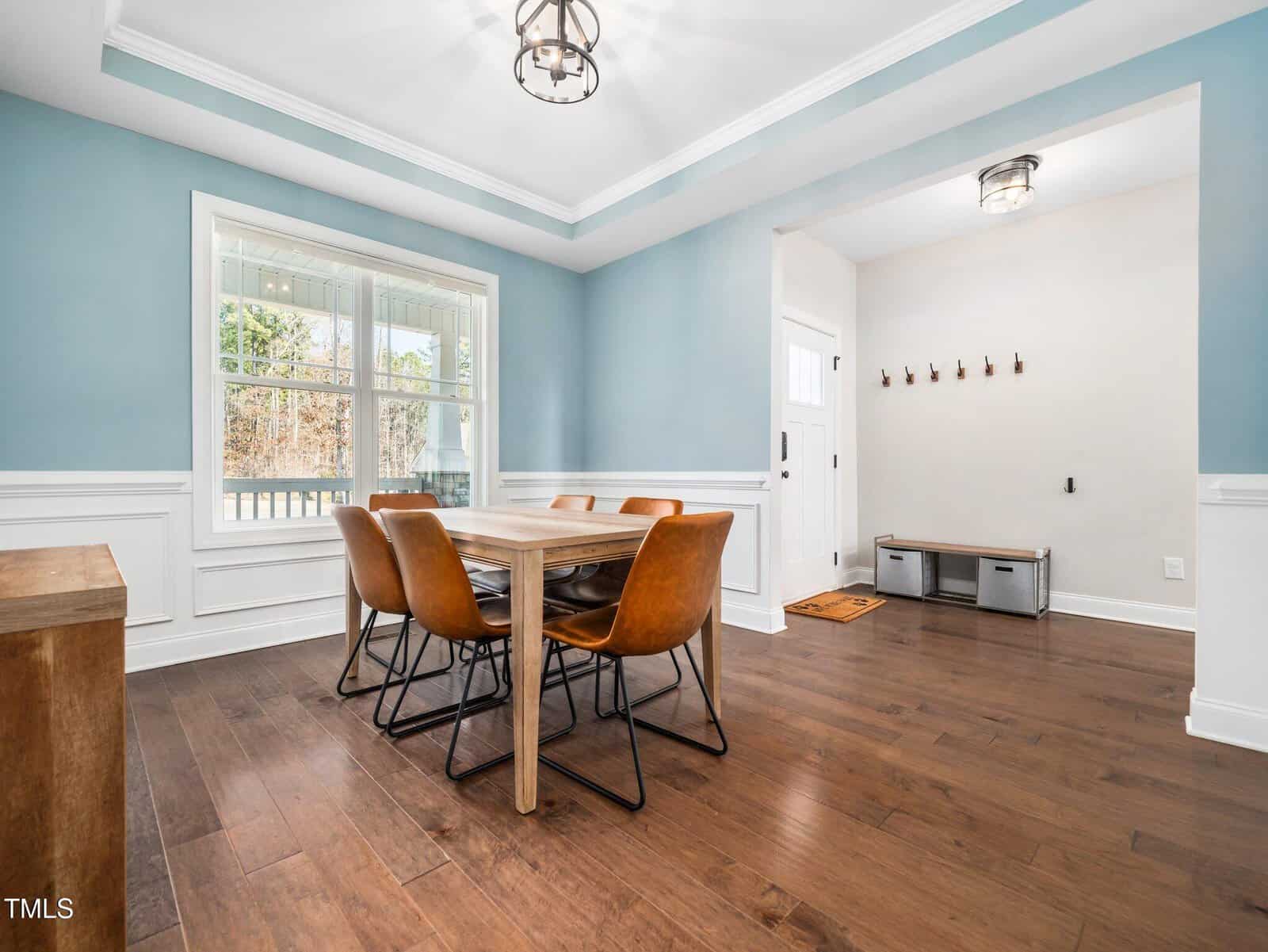 A dining room with light blue walls, white trim, and dark wood flooring features a wooden table with six brown chairs. A window lets in natural light, and an entryway with coat hooks and storage benches is visible.