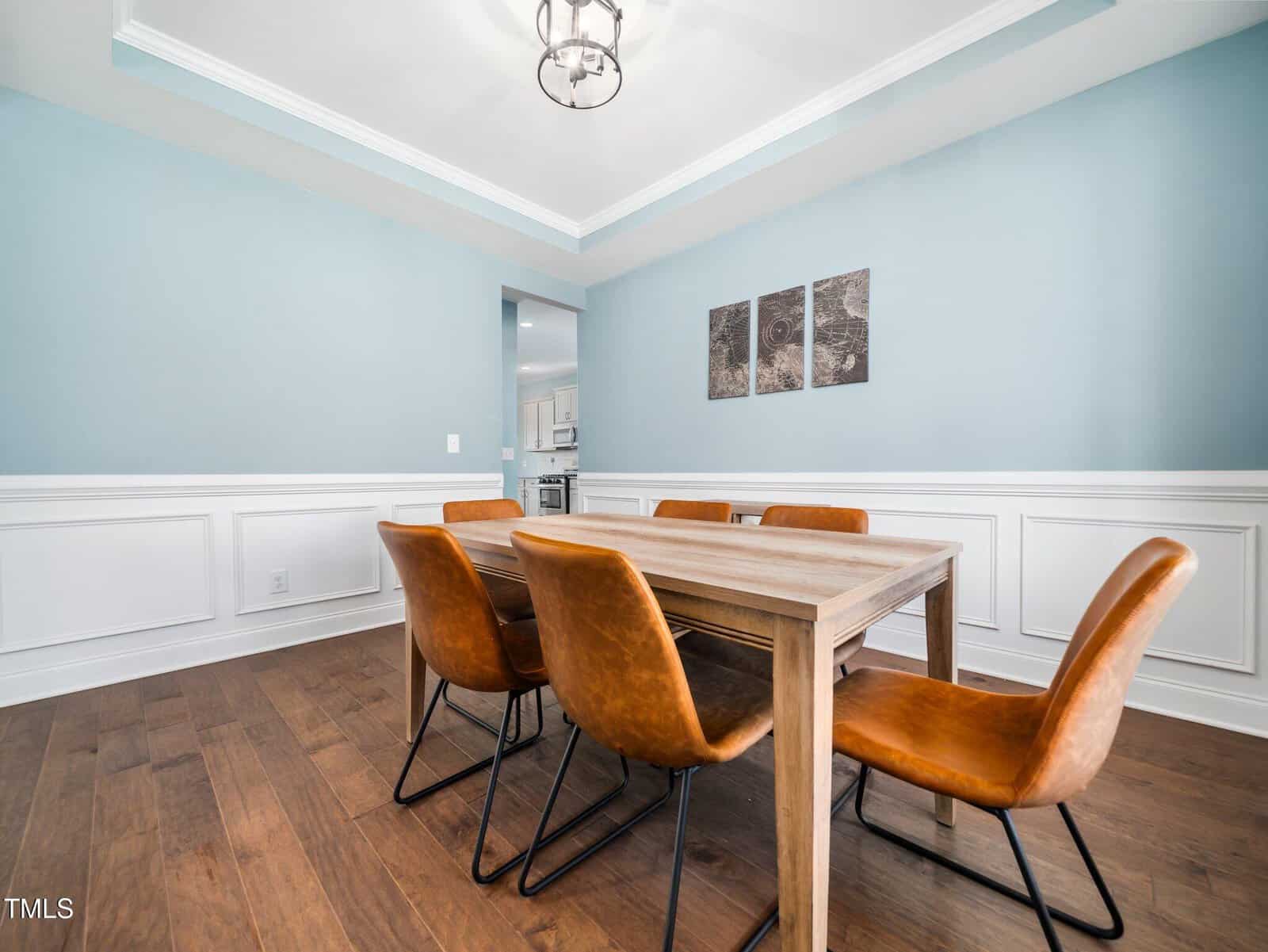 A dining room with a wooden table, six brown leather chairs, light blue walls with white wainscoting, wood flooring, a ceiling light fixture, and two abstract pictures on the wall. An open doorway leads to a kitchen.