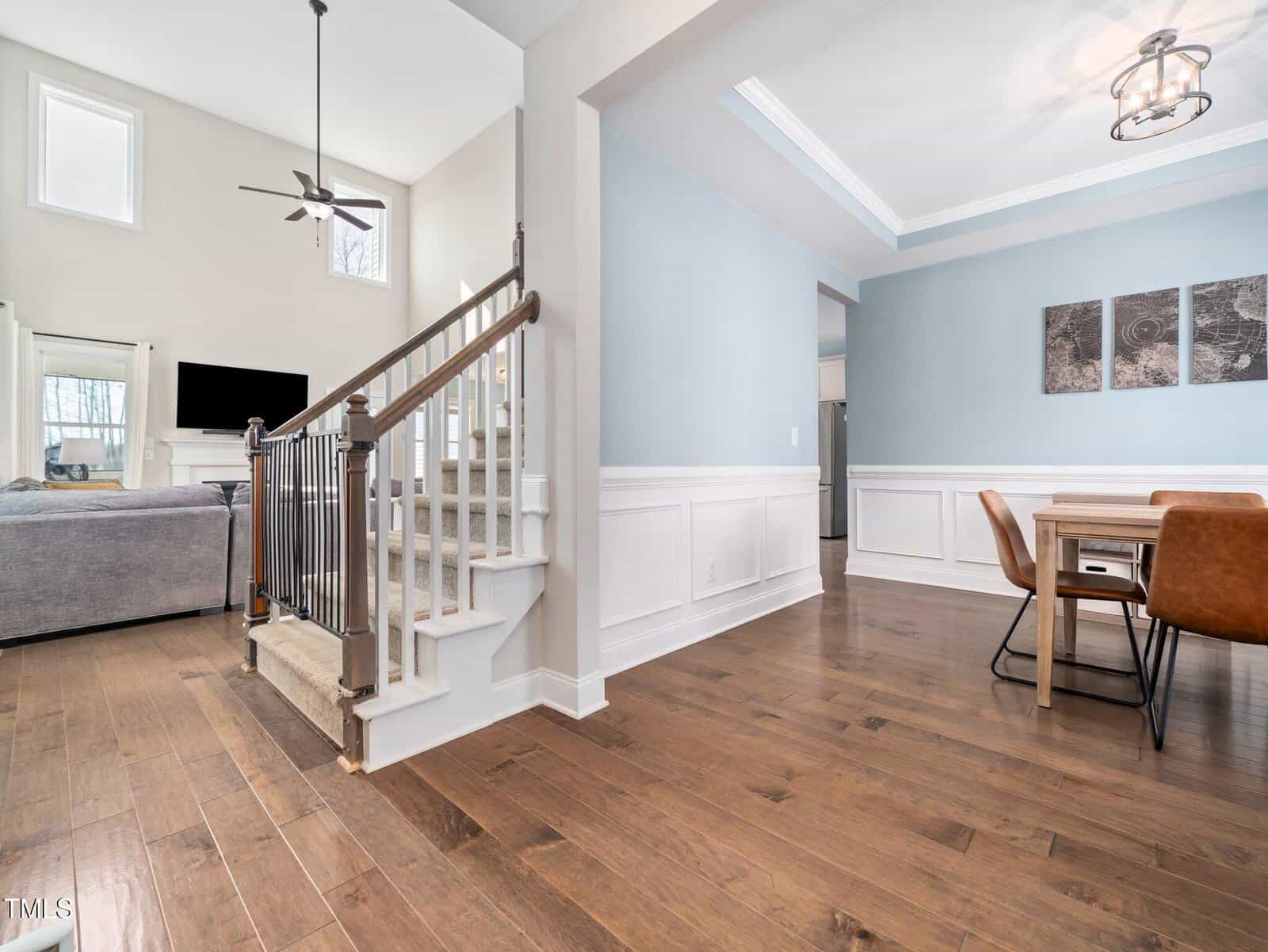 A modern open-concept living and dining area with light blue and white walls, wood flooring, a carpeted staircase with a metal railing, a rectangular dining table, brown chairs, and a ceiling fan.