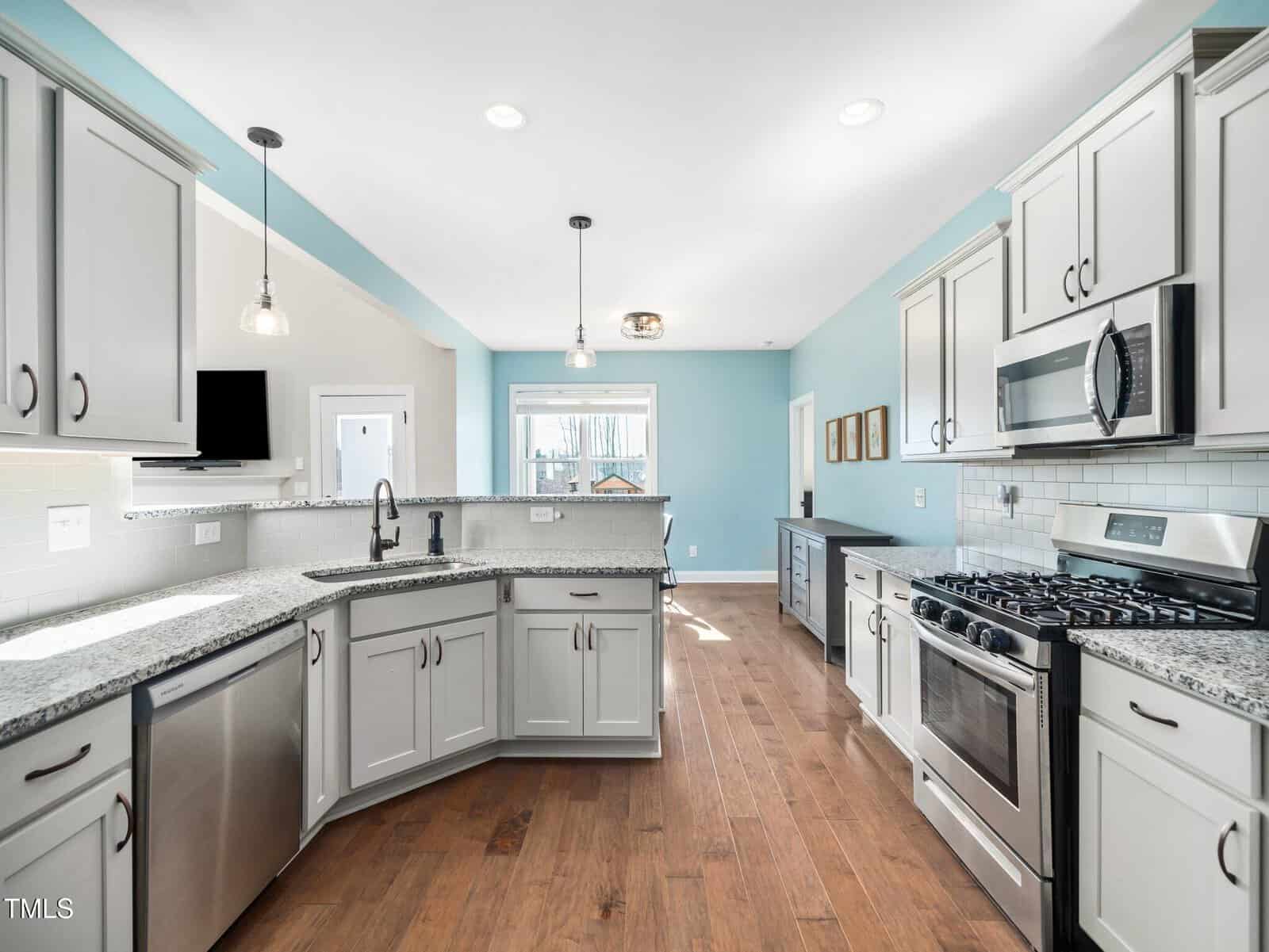 Modern kitchen with light gray cabinets, stainless steel appliances, granite countertops, pendant lights, a farmhouse sink, and light blue walls. Hardwood floors run throughout the space, and natural light fills the room.