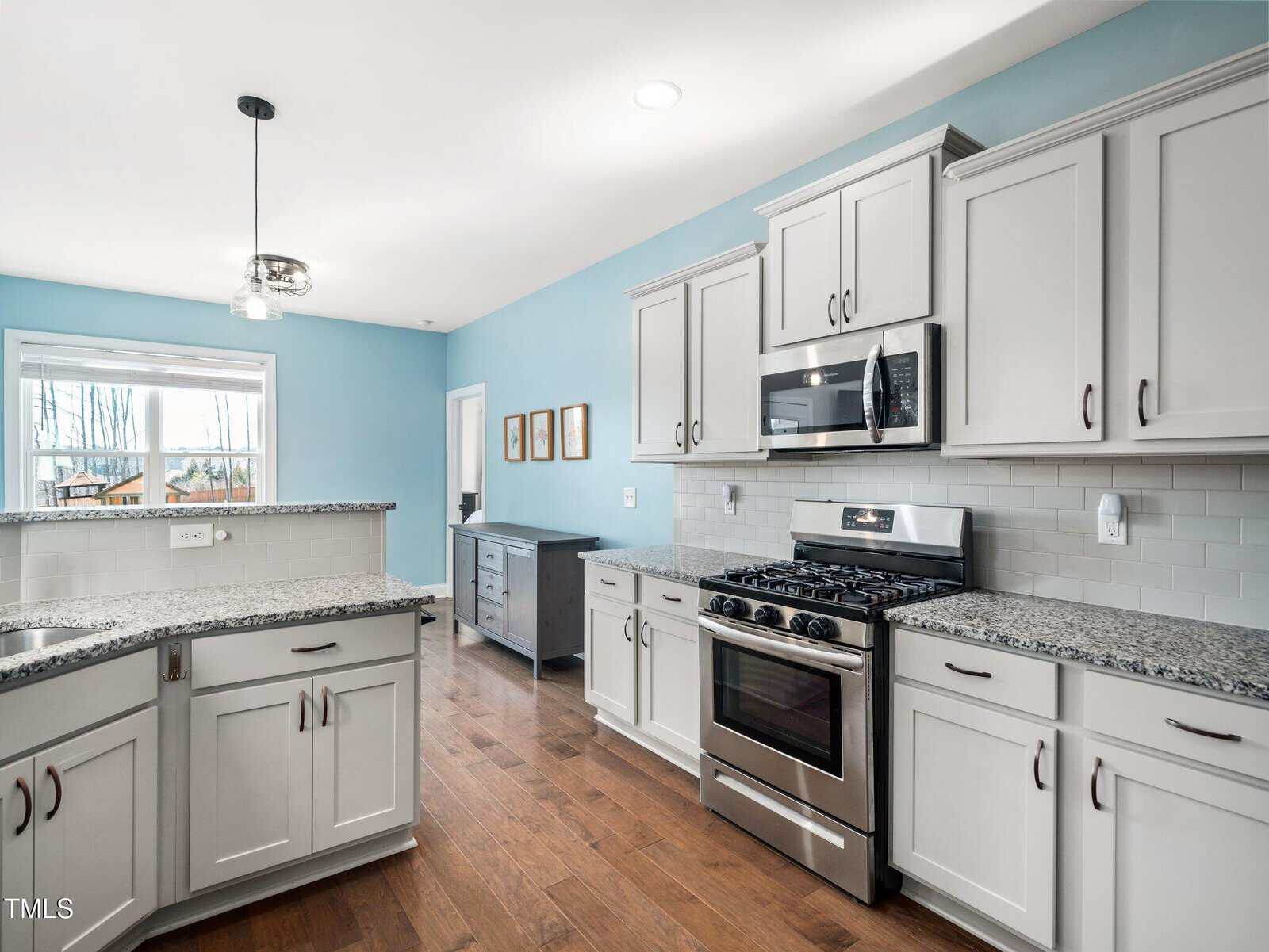 Modern kitchen with light gray cabinets, granite countertops, stainless steel appliances, a gas stove with a microwave above, wood flooring, light blue walls, and a window letting in natural light.
