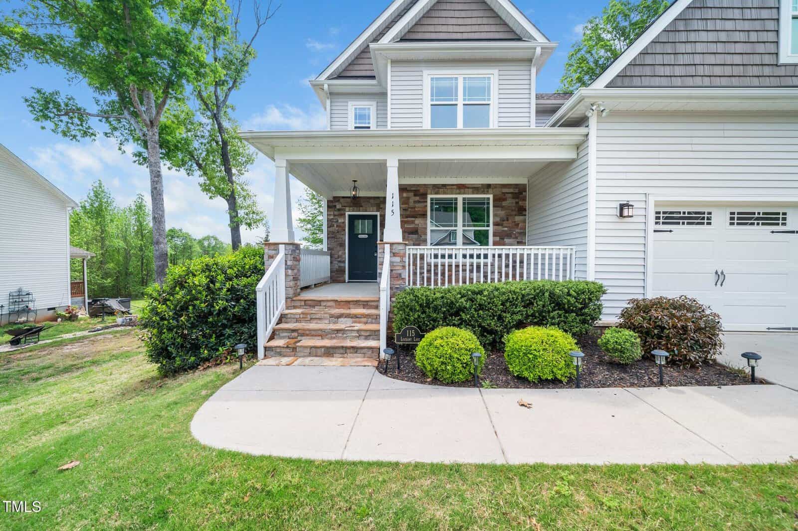 A two-story house with gray siding, stone accents, a black front door, white trim, a covered porch with steps, neatly trimmed bushes, and a concrete walkway leading to the entrance.