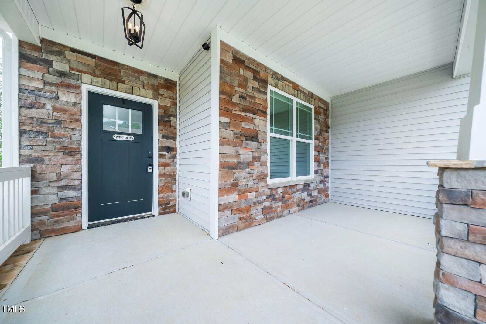 Covered front porch of a house featuring a black front door, stone and siding exterior walls, a large window, and a hanging light fixture. Concrete flooring and white ceiling are also visible.