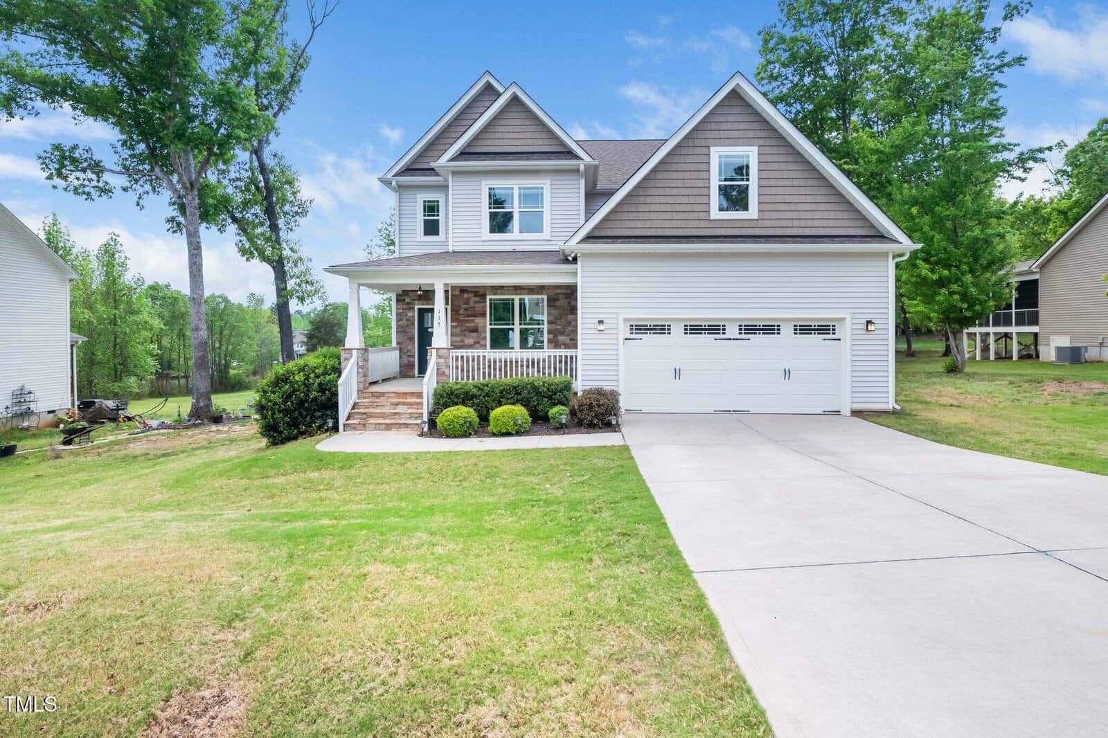 A two-story suburban house with gray siding, white trim, a covered front porch, and an attached two-car garage. The front yard has a sloped lawn with trimmed bushes and a concrete driveway. Trees are in the background.
