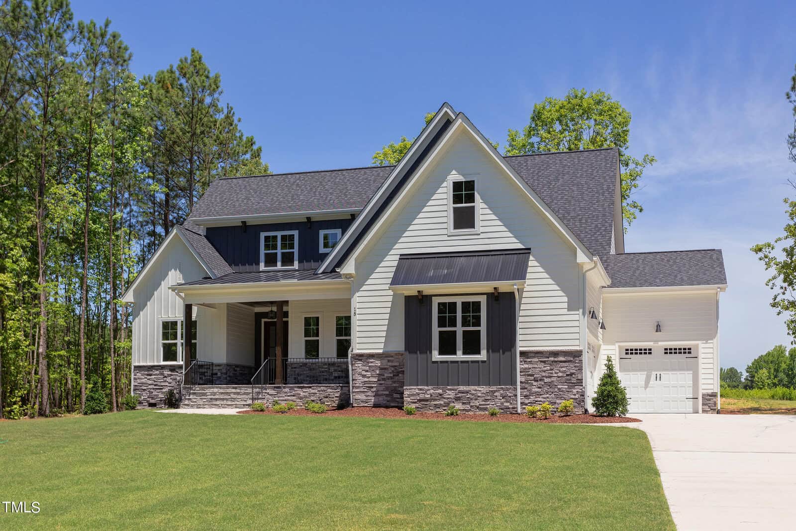 A modern two-story house with white siding, dark gray shingles, stone accents, and a covered front porch, set on a neatly landscaped lawn with trees in the background and a driveway leading to a garage.