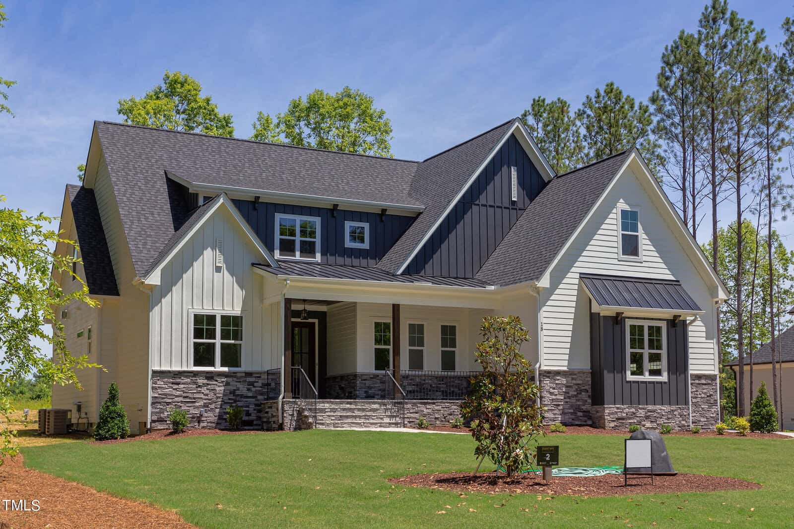 A modern two-story house with white siding, dark roof, and stone accents, featuring large windows, a covered front porch, and a well-maintained lawn with small trees and shrubs under a clear blue sky.