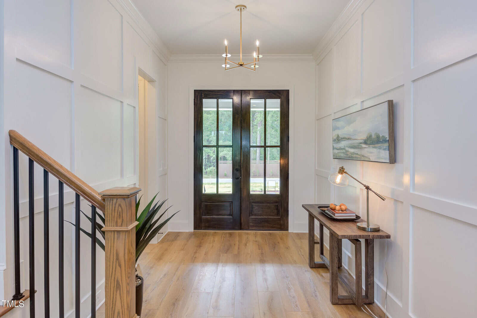 Bright entryway with wooden floors, double dark wood doors with glass panes, a small table with a lamp, art, and bowl. Staircase with black railings on the left, plant below, and chandelier light above.