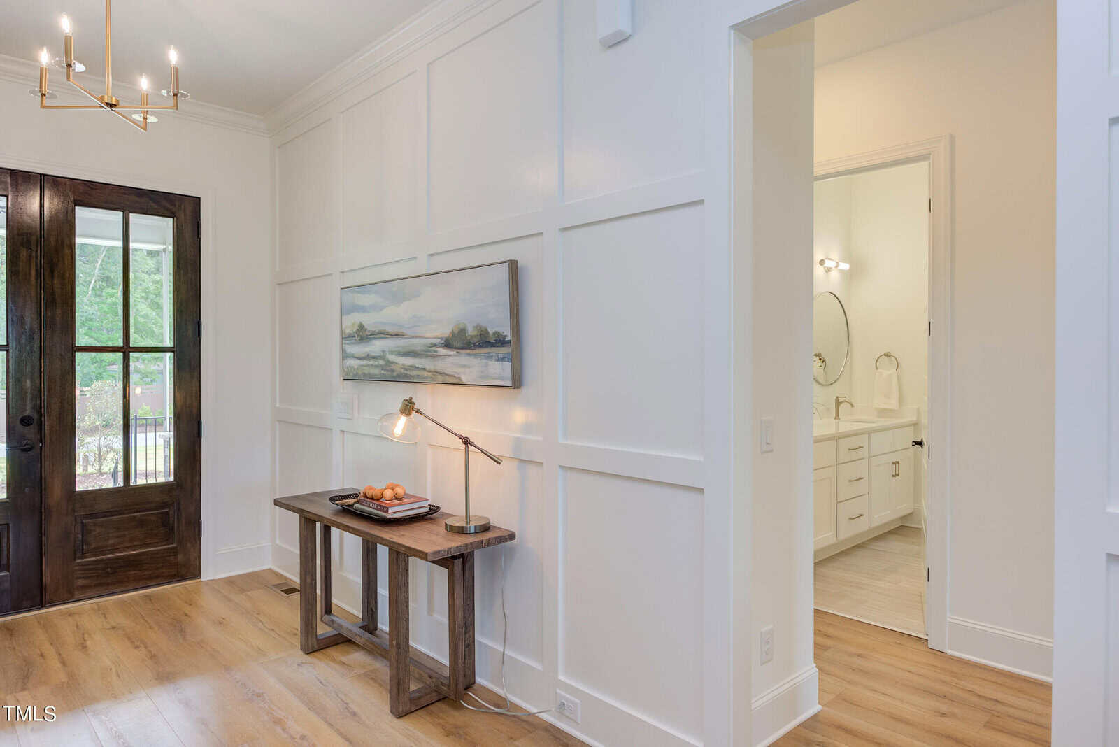 A bright entryway with a wooden bench-style table, lamp, and framed art on a white paneled wall. To the right, an open doorway leads to a bathroom with a vanity and round mirror. Brown double doors are on the left.