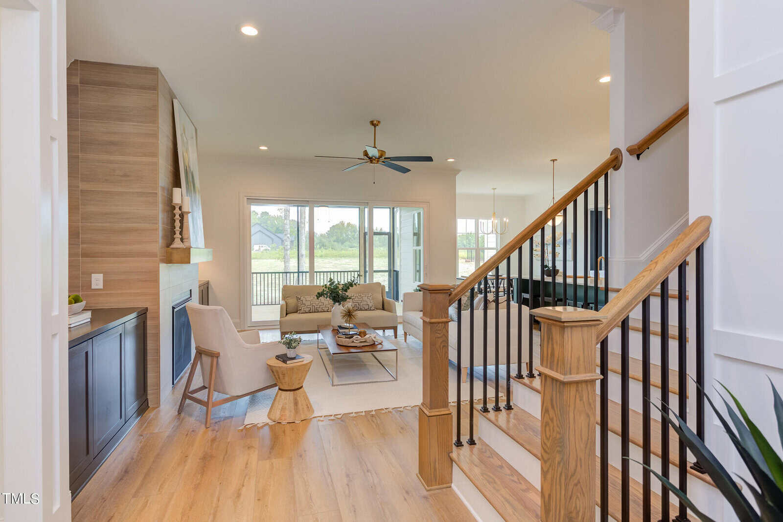 A bright, modern living room with wooden floors, neutral furniture, and a fireplace. Large windows let in natural light, and a wooden staircase with black railings is in the foreground. A ceiling fan hangs above the seating area.