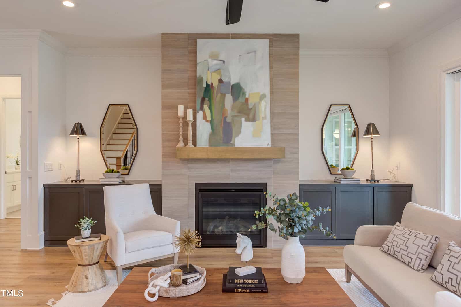 A modern living room with a fireplace, abstract art above the mantel, two mirrors and lamps on side cabinets, a white armchair, light sofa, coffee table with books and decor, and hardwood floors.