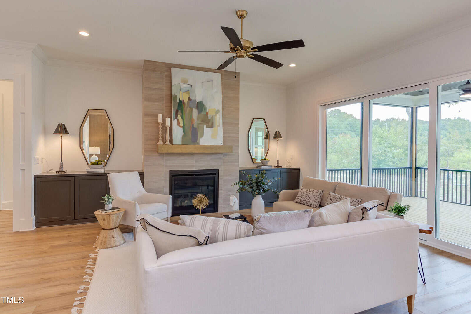 A modern living room with a white sofa, armchair, central coffee table, and a fireplace with abstract art above it. Large windows provide a view of greenery outside, and there are two mirrors and lamps on either side of the fireplace.