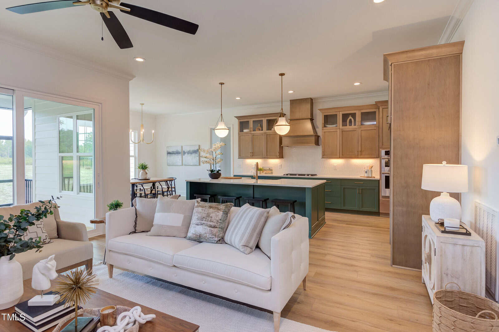 A bright, modern open-concept living room and kitchen with light wood floors, a cream sofa with throw pillows, a kitchen island with green cabinetry, pendant lights, and large windows letting in natural light.