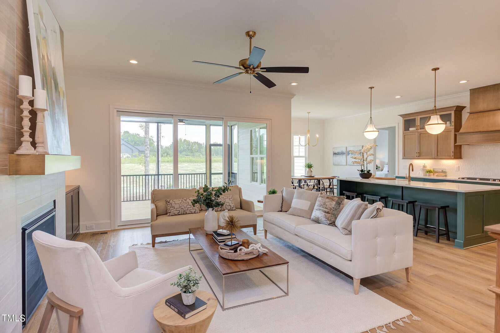 Bright, modern living room with neutral furniture, a fireplace, wooden floors, and large windows. The space opens to a dining area with pendant lights and a green kitchen island, overlooking a scenic outdoor view.