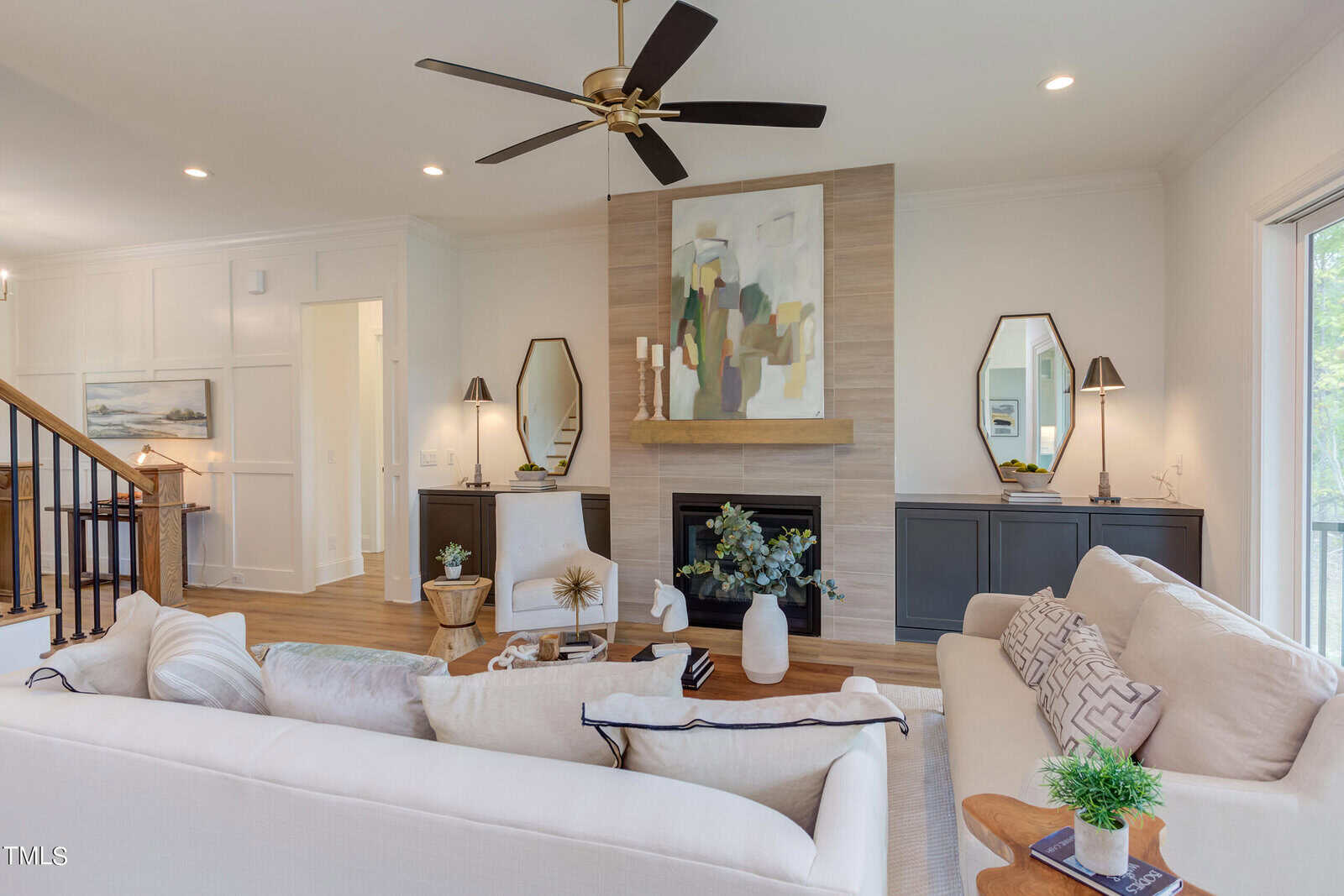 Bright living room with a white sofa, modern art above a fireplace, a ceiling fan, two armchairs, built-in black cabinets with mirrors, and large windows letting in natural light.