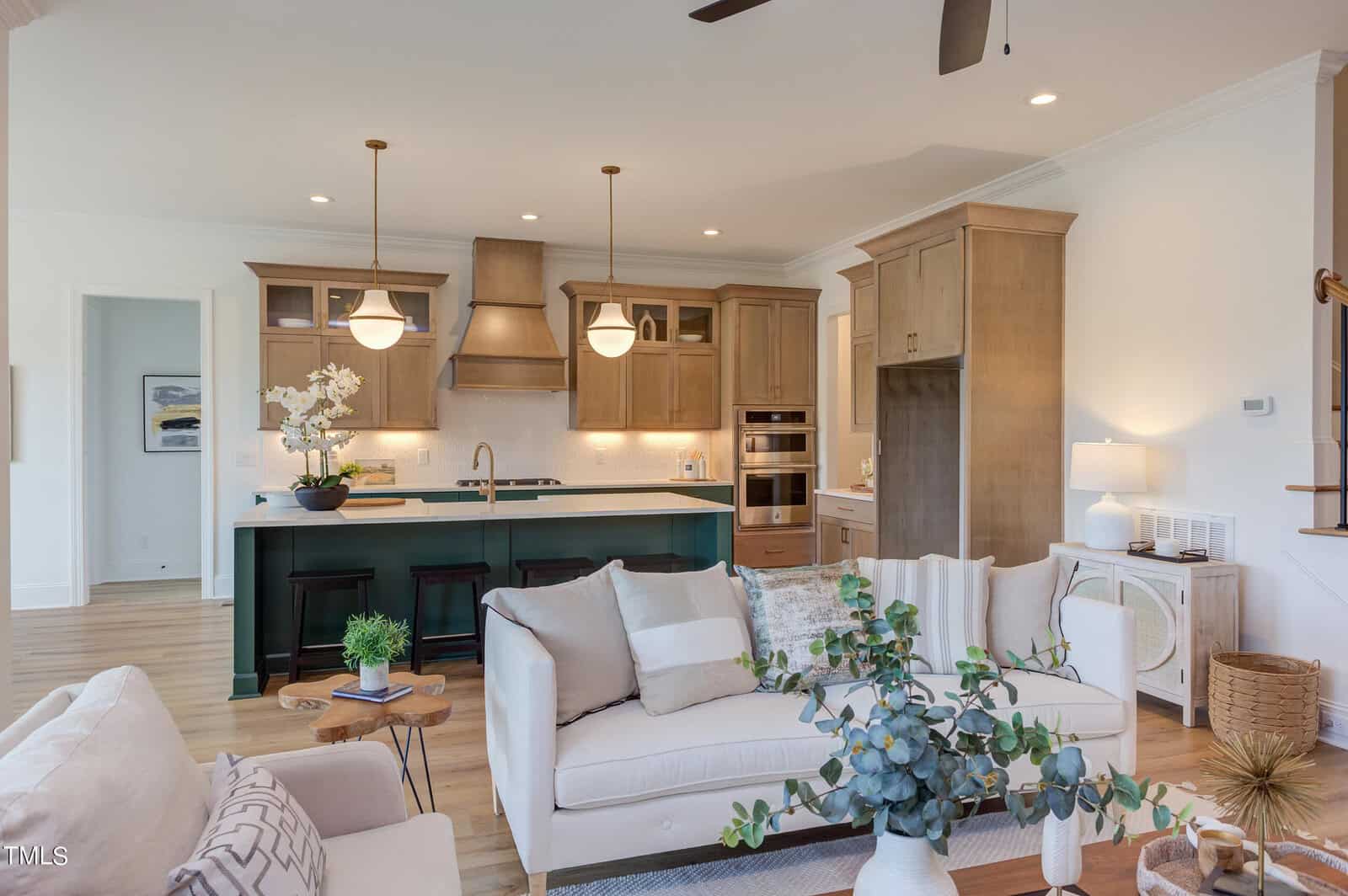 Modern open kitchen and living room with wood cabinets, green island, pendant lights, beige sofa, armchairs, and plants. Light-filled, cozy, and stylish space with minimal decor and a welcoming atmosphere.