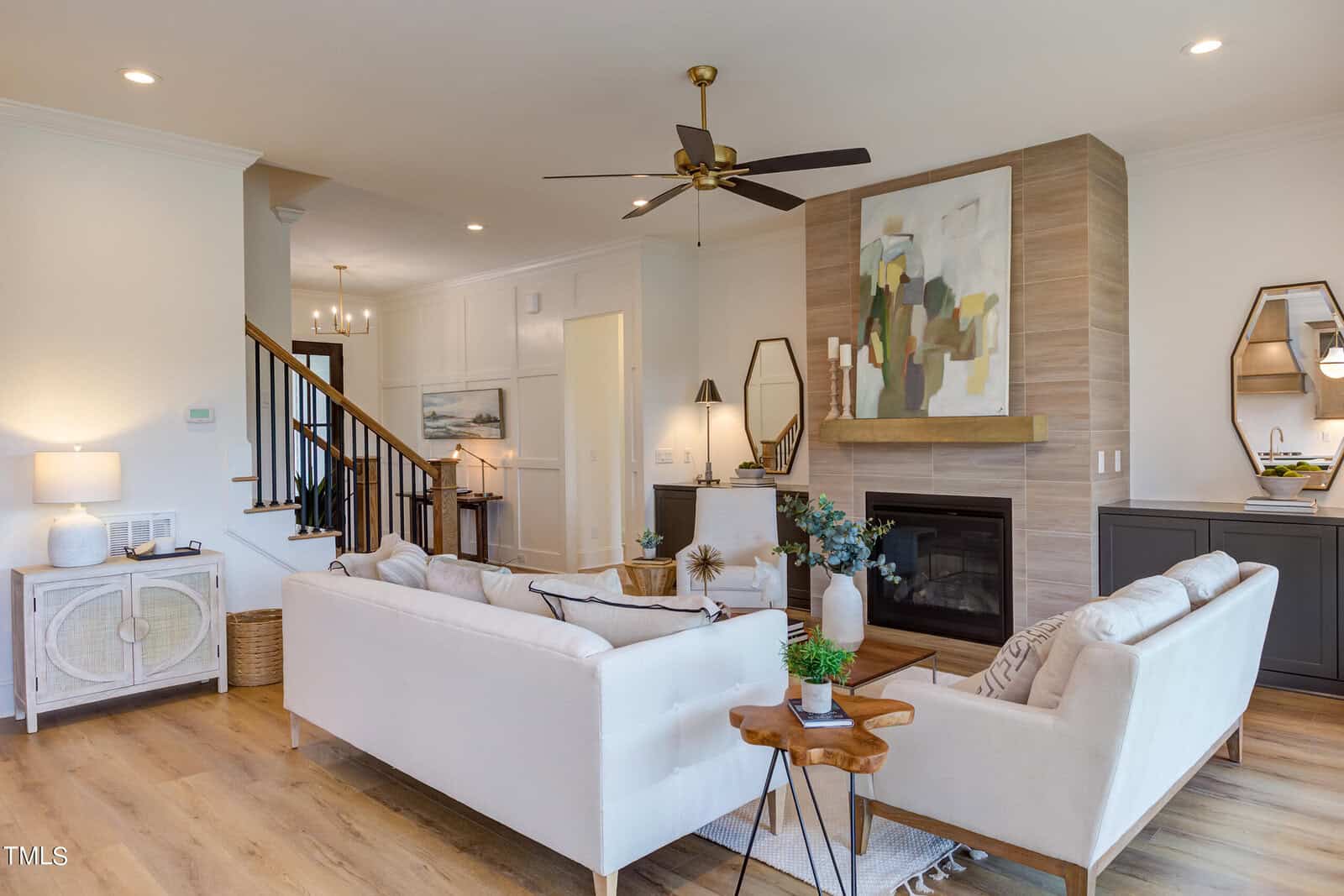 Modern living room with light sofas, abstract art above a fireplace, wood flooring, ceiling fan, and decorative mirrors on side tables; stairs and hallway are visible in the background.