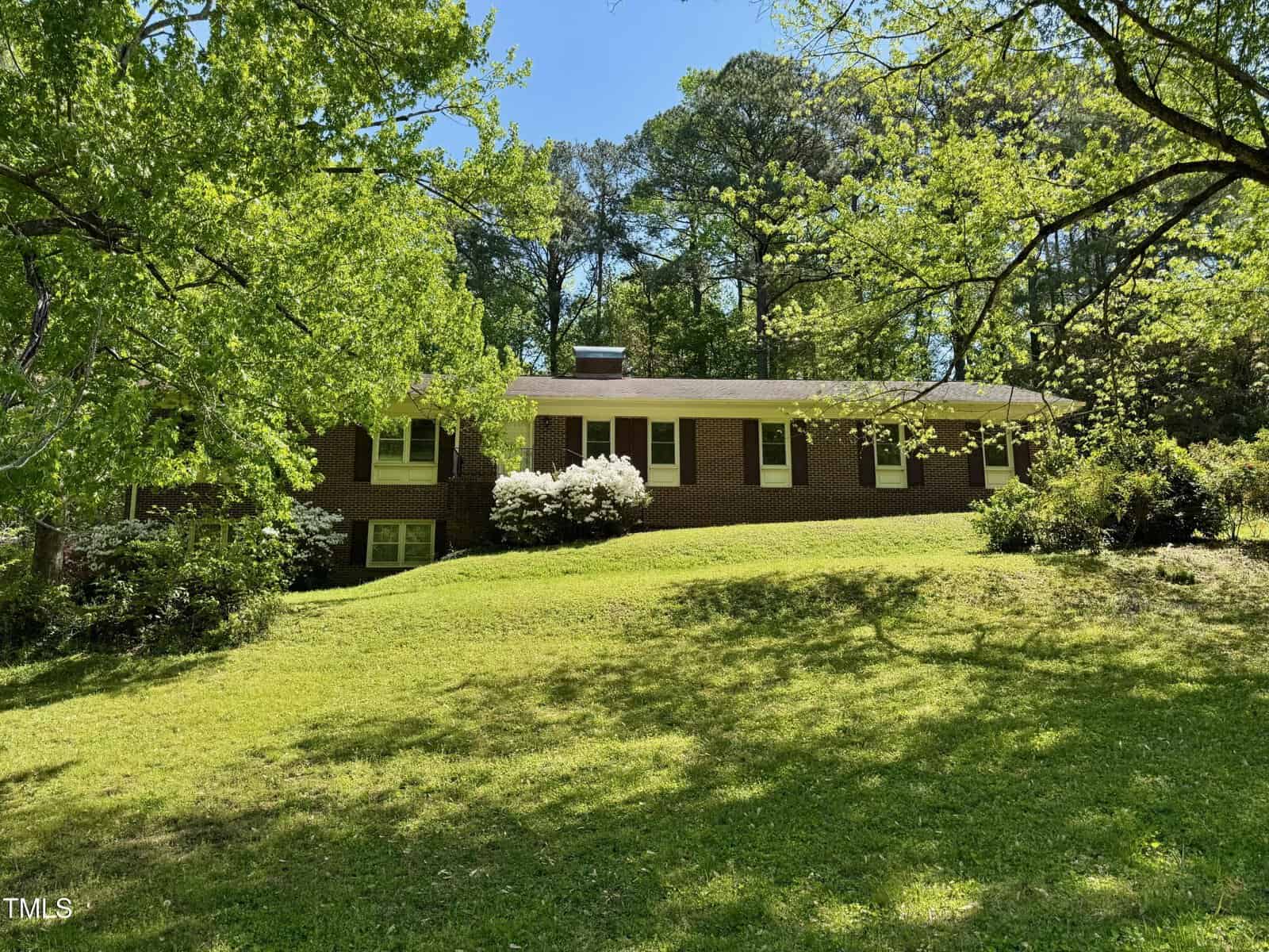 A single-story brick house with white trim sits on a grassy hill at 2118 N Woodland Road in Henderson, surrounded by green trees and bushes under a clear blue sky.