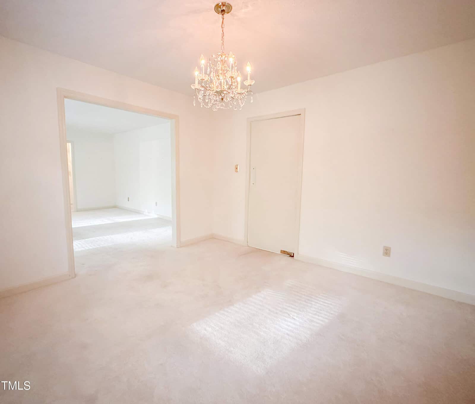 A bright, empty room with off-white walls and carpet, featuring a chandelier, an open doorway leading to another room, and a closed sliding door on one wall. Sunlight streams in through a window out of view.