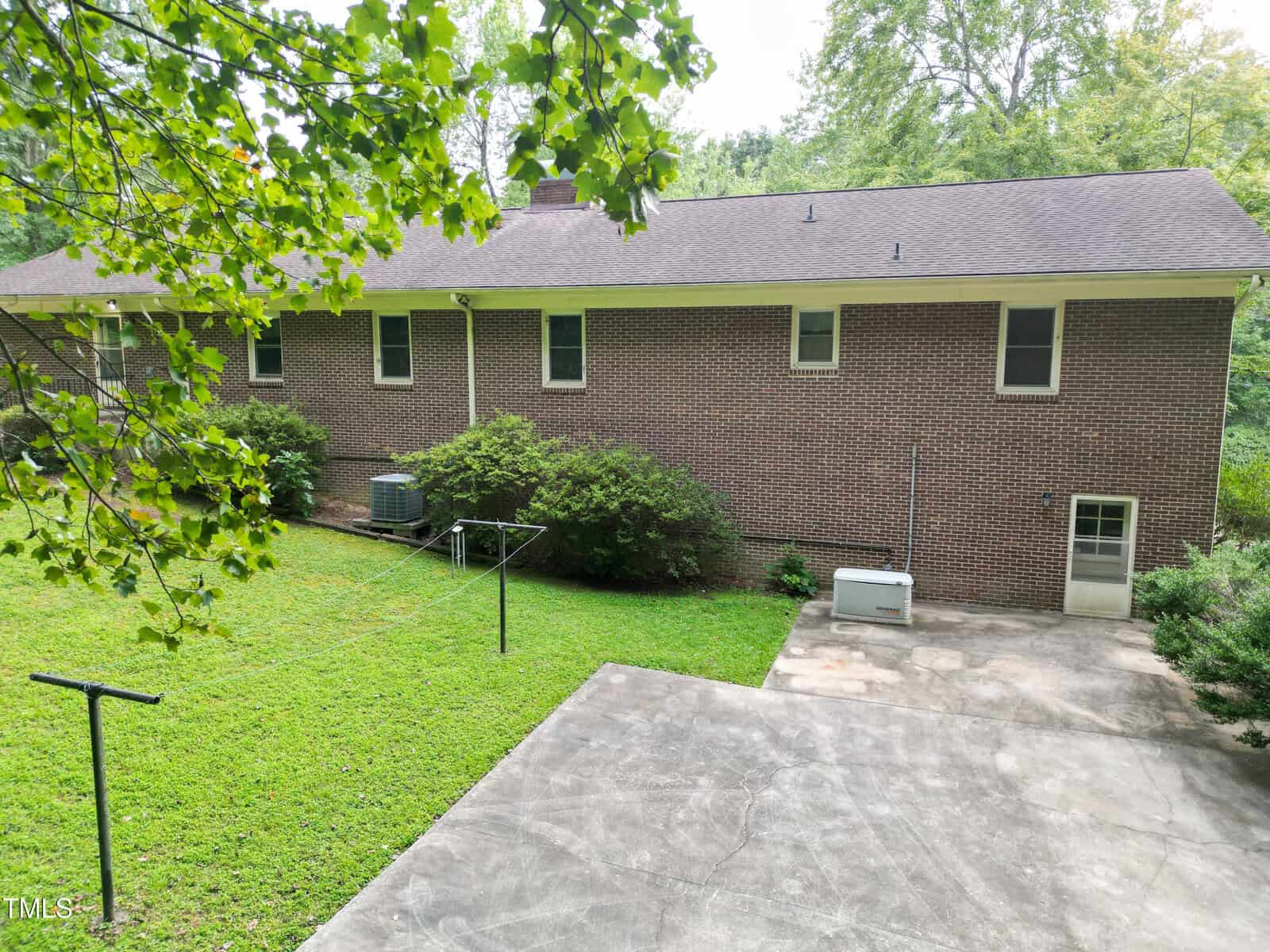 A brick house with a shingled roof, viewed from the backyard, featuring a concrete patio, a lawn with clothesline poles, air conditioning units, and a backup generator. Trees and bushes surround the yard.