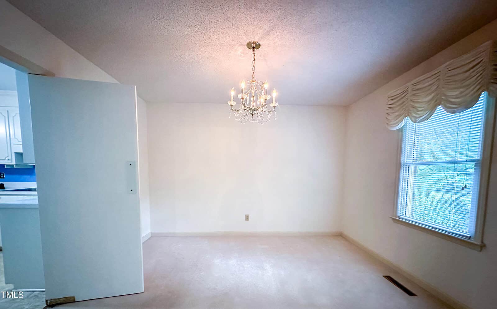 Empty room with cream-colored carpet, white walls, and a chandelier hanging from the ceiling. There is a large window with drapes on the right and an open door leading to a kitchen on the left.