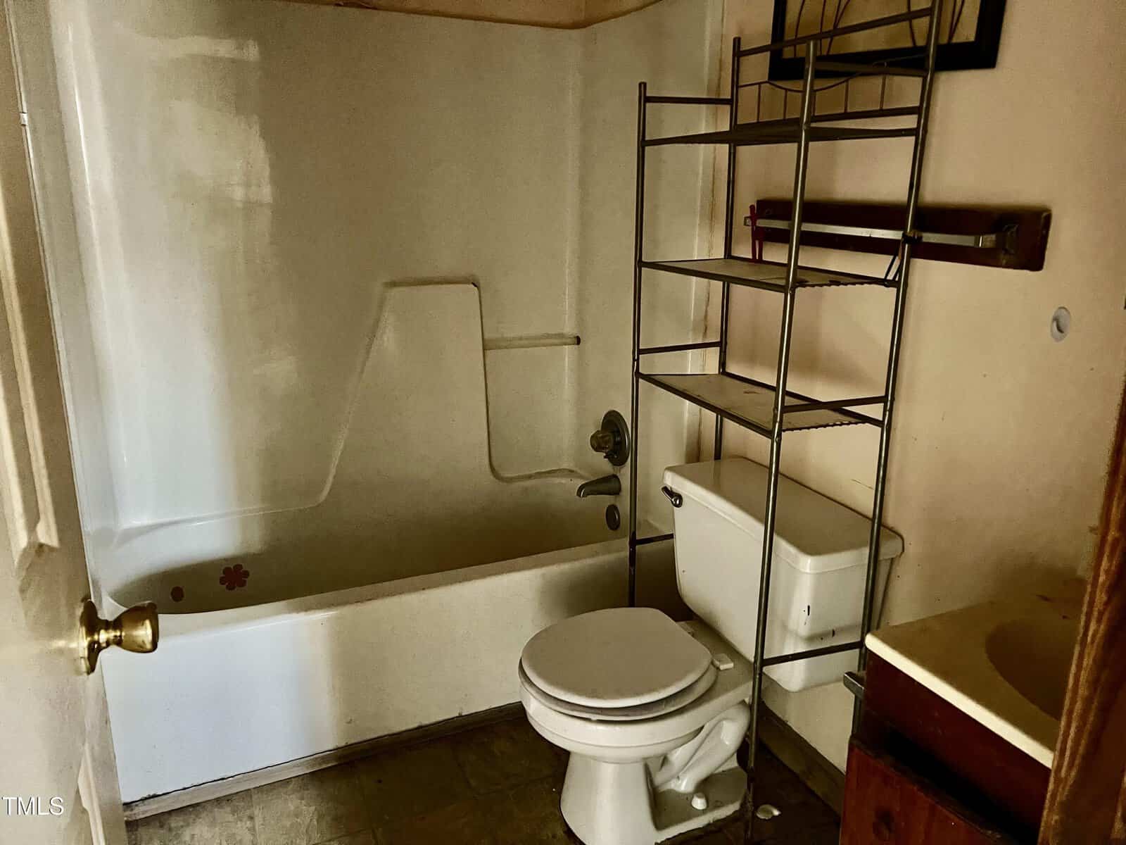A small, dimly lit bathroom with a white bathtub, a toilet, and a metal shelving unit above the toilet. The floor appears to be tiled, and there's a sink with a brown cabinet to the right. A white door is partially open.